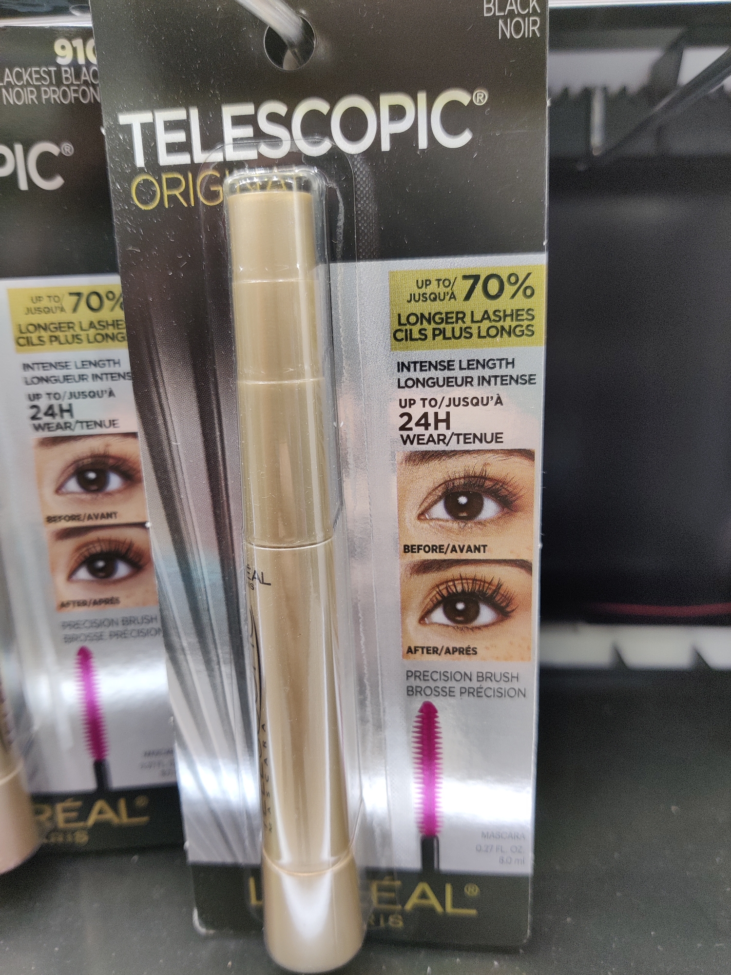 The image shows a close-up of a L'Oréal Telescopic Original mascara product, likely displayed in a retail setting. The packaging is primarily black with white and gold lettering. The mascara tube itself is a golden color, and it is enclosed in clear plastic packaging. On the left side of the packaging, there are before and after images of eyes demonstrating the mascara's lengthening effect. Text on the packaging highlights "Up to 70% Longer Lashes" and "24H Wear/Tenue." The product is labeled "Black Noir." In the background, shelves and other retail items are visible, with a dark, blurry backdrop. The lighting suggests an indoor retail environment. This product appears to be for sale in a store in Mechanicsburg, United States of America.