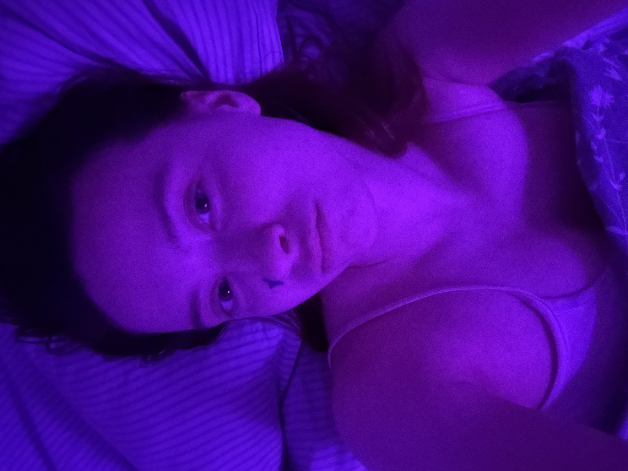 A woman is lying in bed under a purple light. She is wearing a pink tank top. The sheets are striped and there is a floral patterned blanket near her. The image does not provide enough information to identify a specific location in Прикамский, Russia.