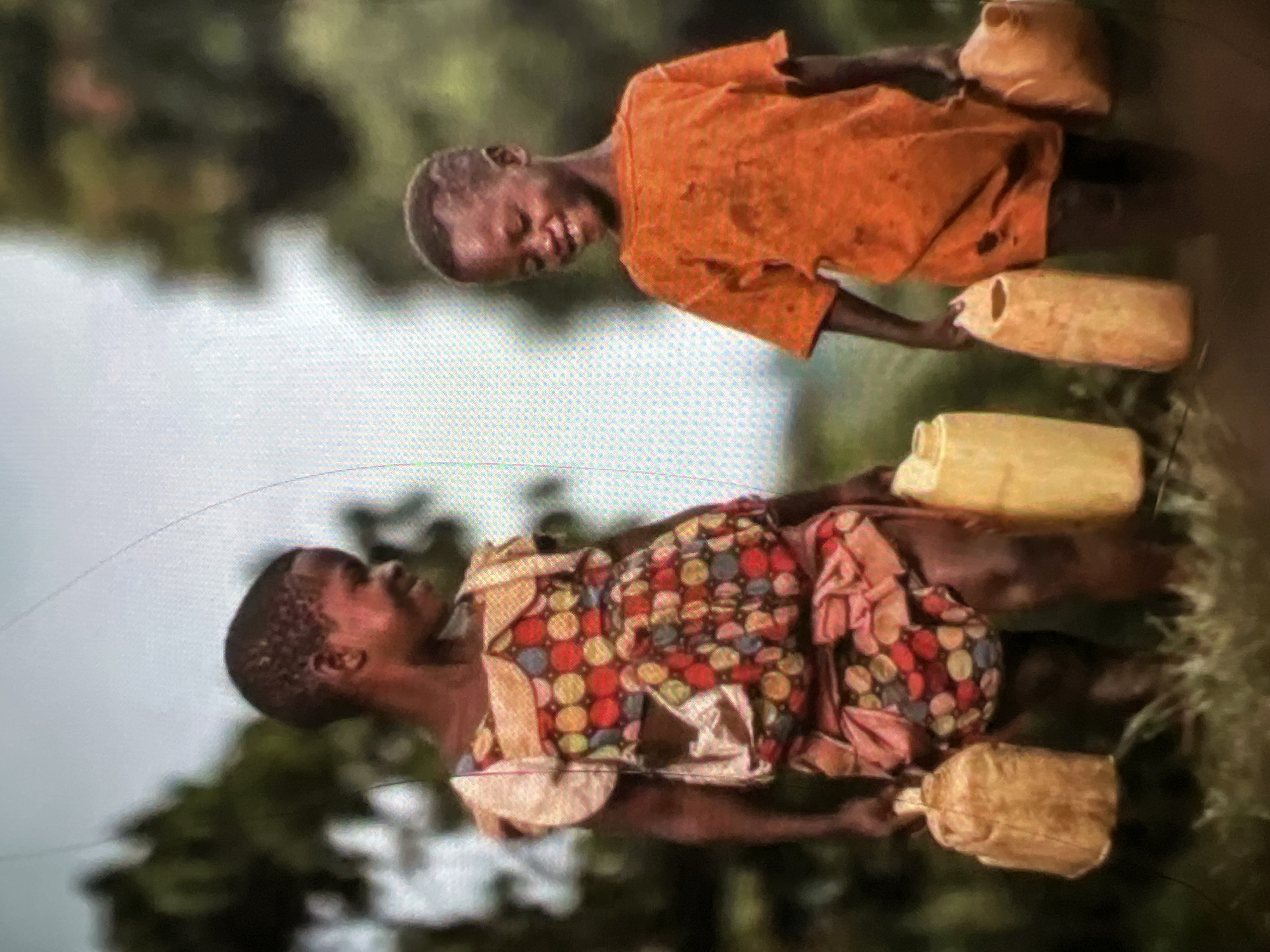 Two young children, a girl and a boy, stand in a grassy, outdoor environment. The girl on the left wears a vibrant, multi-colored dress adorned with a pattern of red, yellow, and blue circles. She carries a light brown, cylindrical jug in her left hand and another identical jug in her right. The boy on the right is dressed in a faded orange t-shirt and carries a light brown, tall, narrow jug in his right hand, and a similar jug in his left. The children appear to be interacting, with both looking down and smiling. The background is softly blurred, suggesting trees and foliage. The scene evokes a sense of shared task and youthful resilience. The location is not identifiable as Lagos, Nigeria, or any specific city.