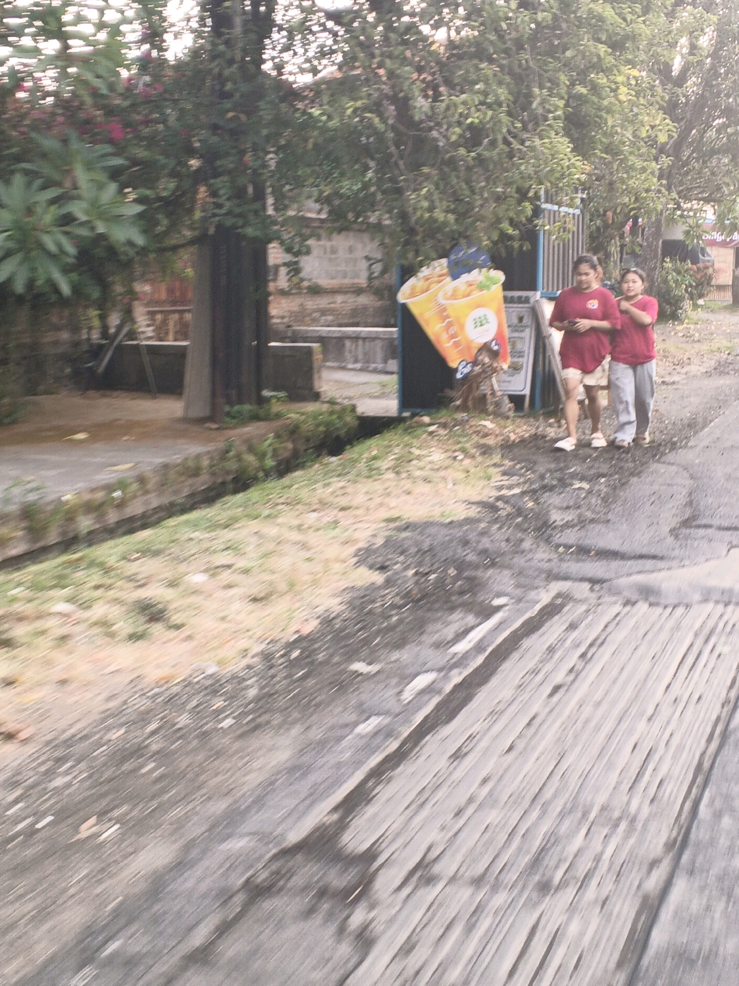 Two women in red shirts and one wearing gray pants and the other white shorts are walking on a street in Sukawati, Indonesia. A large cardboard cutout of a drink is to their left, leaning against a fence. Trees and buildings are visible in the background. The street is paved and appears to be wet in some areas. The scene is captured from a low angle, suggesting the photo was taken from a moving vehicle.