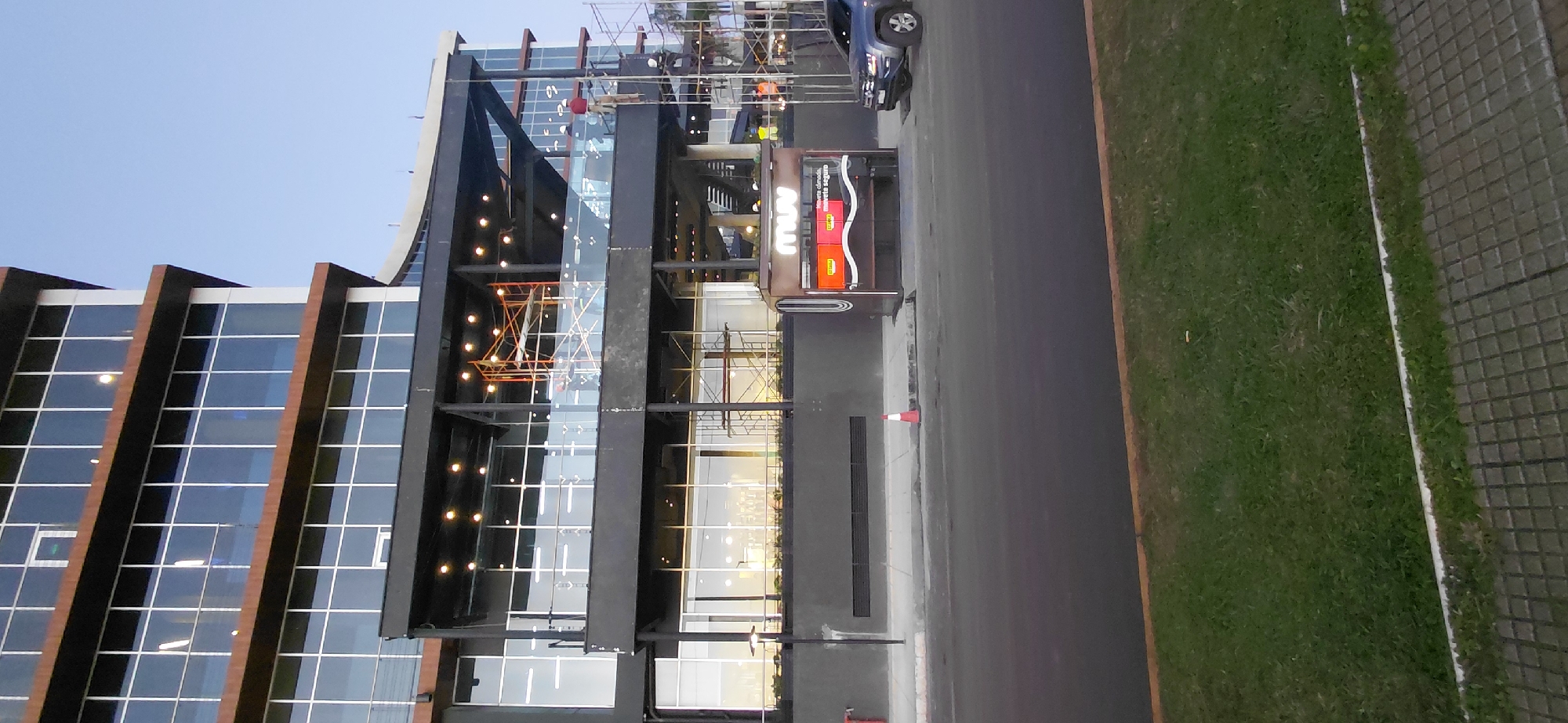 This image captures the exterior of a multi-story building in Encarnación, Paraguay. The building features a modern design with extensive glass facades and dark-colored structural elements. Scaffolding is visible on one side of the building, indicating ongoing construction or renovation work. Several construction workers in hard hats and safety vests are present on the scaffolding and on a balcony area. In the foreground, a dark-colored car is parked near a small kiosk or bus stop structure. A traffic cone is placed on the sidewalk near the kiosk. The scene is set against a clear blue sky, with a strip of grass and a paved walkway separating the building from the street. The overall impression is a scene of urban development or maintenance.