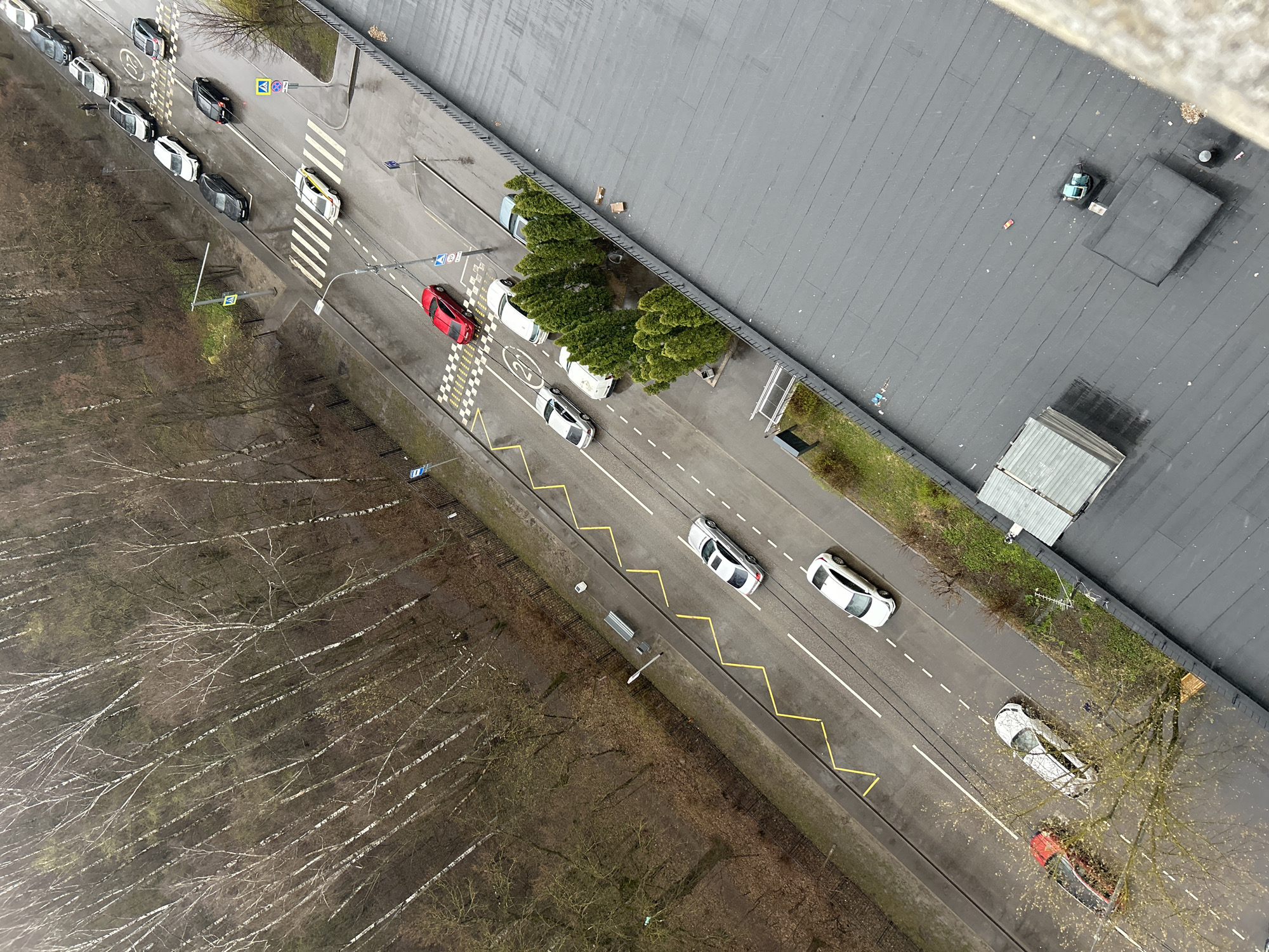 The image is an aerial view of a street in Moscow, Russia. The street is lined with trees and buildings. There are cars parked on both sides of the street. The trees are mostly bare, suggesting it is autumn or winter. The road features painted lines, a crosswalk, and speed bumps. The rooftops of buildings, one appears to be a large commercial or industrial structure, are seen on the right side of the image. A forest of bare trees is visible on the left. The lighting is overcast.