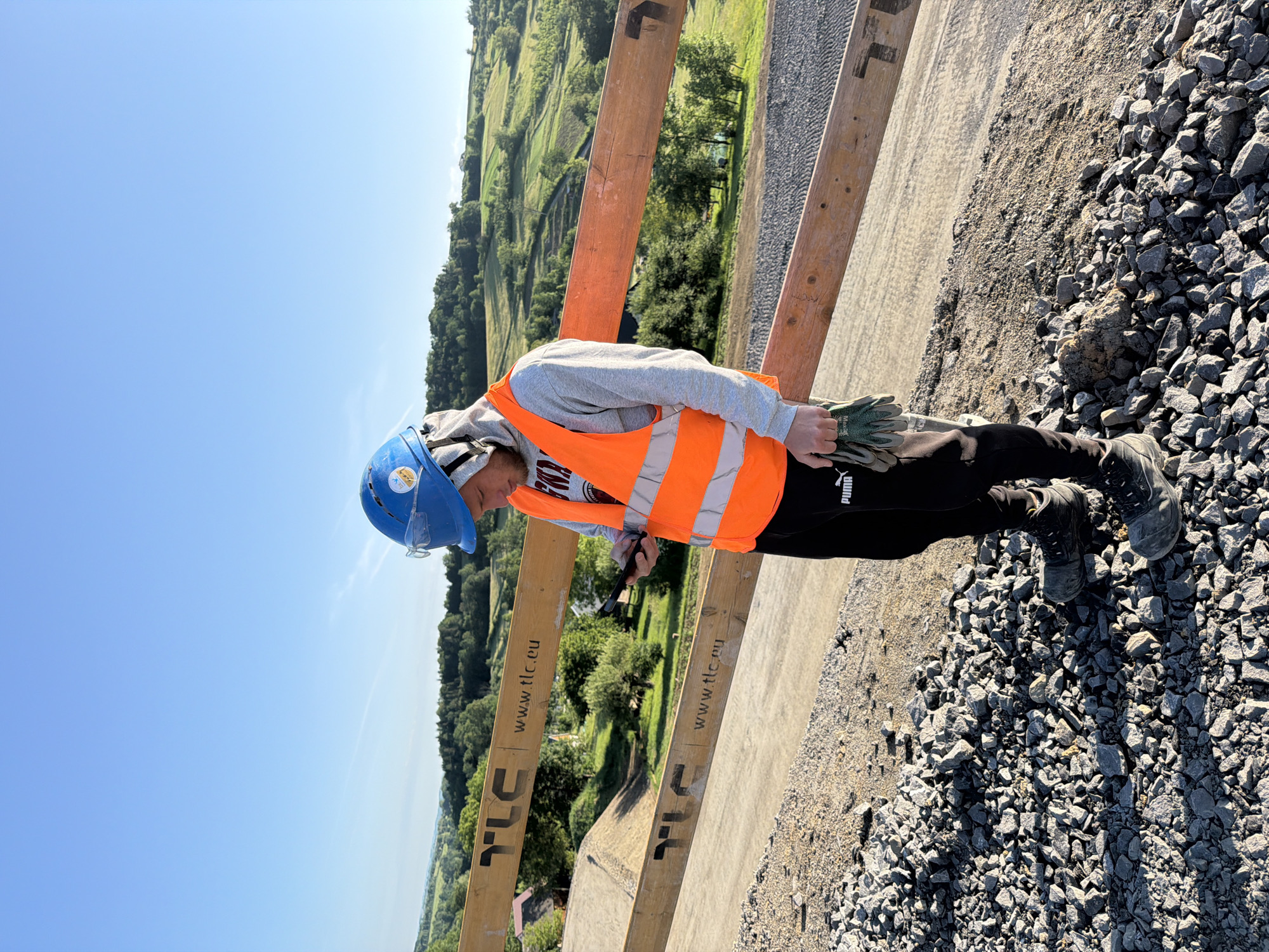 A construction worker, wearing a blue hard hat with safety glasses, an orange reflective vest over a gray hooded sweatshirt, and black Puma sweatpants, stands on a gravel-covered embankment. He is holding a black smartphone in his right hand and a pair of work gloves in his left. The worker is looking down at the phone.
Behind him, a wooden barrier with the "TLC" logo and "www.tlc.eu" printed on it runs horizontally across the scene. Beyond the barrier, a lush, green, hilly landscape stretches under a clear blue sky. Several houses and trees are visible nestled within the rolling hills, suggesting a rural or semi-rural setting. The area appears to be under construction, indicated by the gravel and the unfinished embankment. The location is identified as Stróża, Poland.