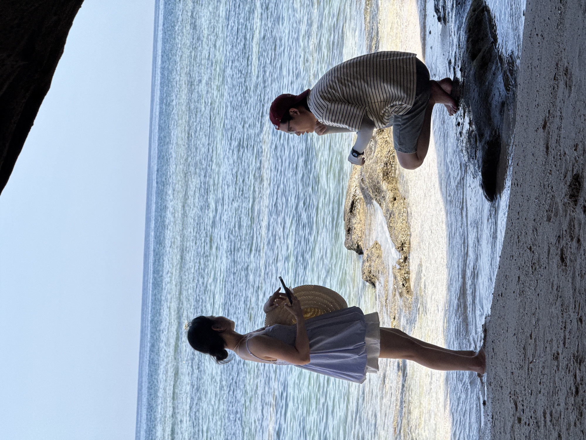 Two individuals are positioned near the shoreline on Lý Sơn Island, Vietnam. A woman stands on the sandy beach, holding a straw hat and a smartphone. She is wearing a light blue dress over white shorts. Facing her, a man is crouched on a rock in the shallow water. He is wearing a striped t-shirt, shorts, a red cap, and glasses. The ocean stretches out behind them, with a clear sky above. The image captures a moment of interaction or observation between the two people in this coastal setting.