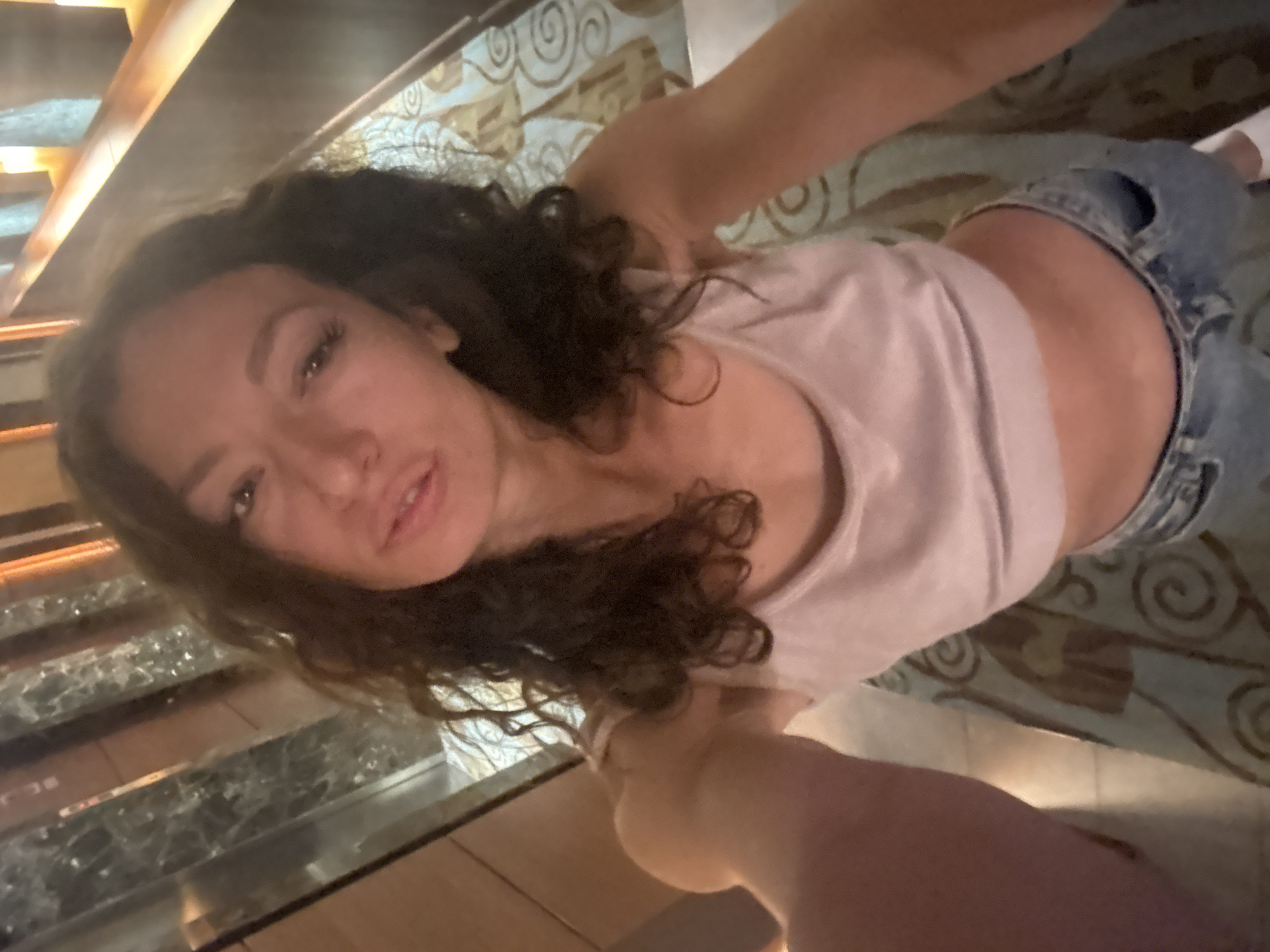 A woman with curly dark hair is looking at the camera. She is wearing a light-colored tank top and blue denim shorts. The background includes a staircase and a carpet with a circular pattern. The location is potentially Đà Nẵng, Vietnam, but there is no explicit visual information in the image to confirm this.