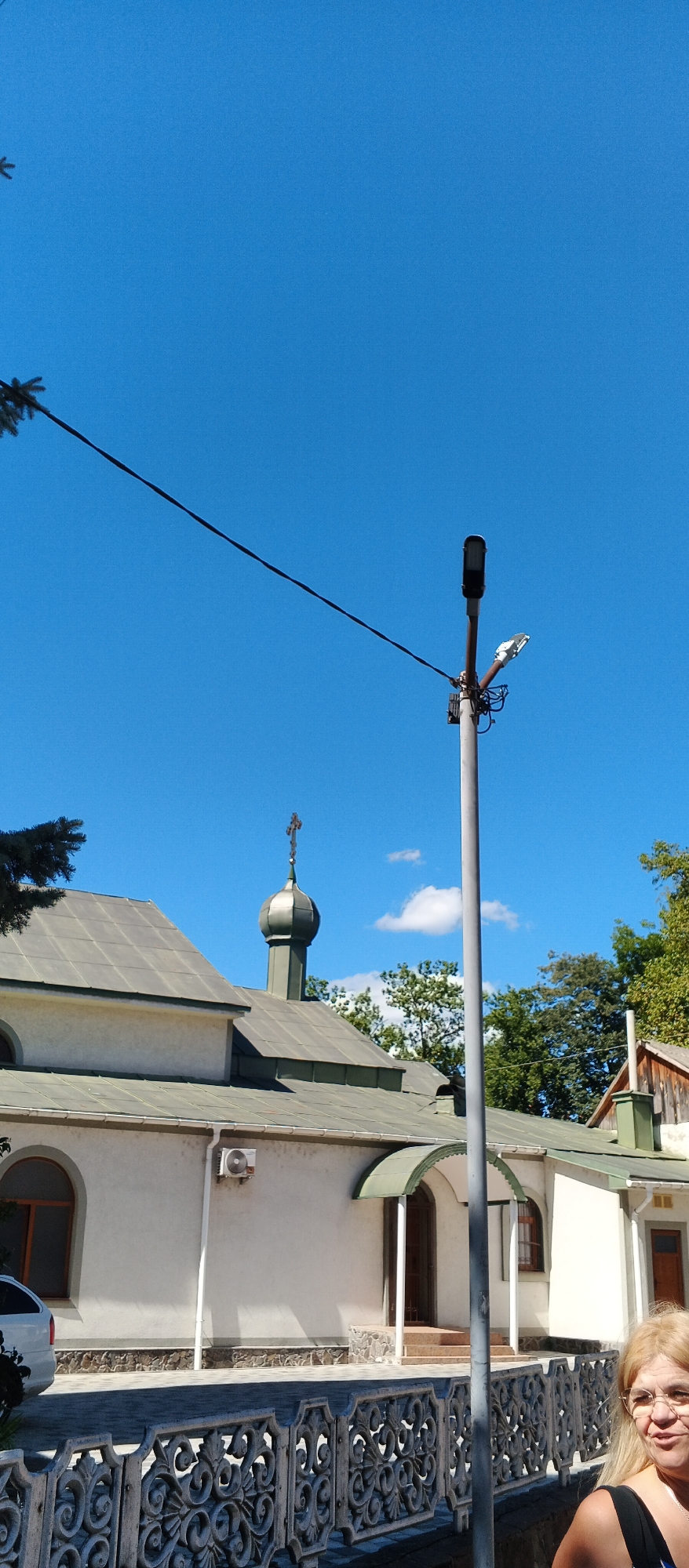 The image shows a frontal view of a white building with a green roof, possibly a church, located in Mukachevo, Ukraine. A prominent feature is a dome topped with a cross, characteristic of Eastern Orthodox architecture. In the foreground, a decorative metal fence lines the property. To the right of the building, a street lamp post stands tall, with two lights mounted on it. Behind the building, lush green trees form a dense backdrop against a clear blue sky dotted with a few white clouds. The lower right corner of the image shows the upper body of a blonde woman with glasses, looking to her right and smiling. The scene appears to be a sunny day, suggesting it was taken outdoors during daylight hours.