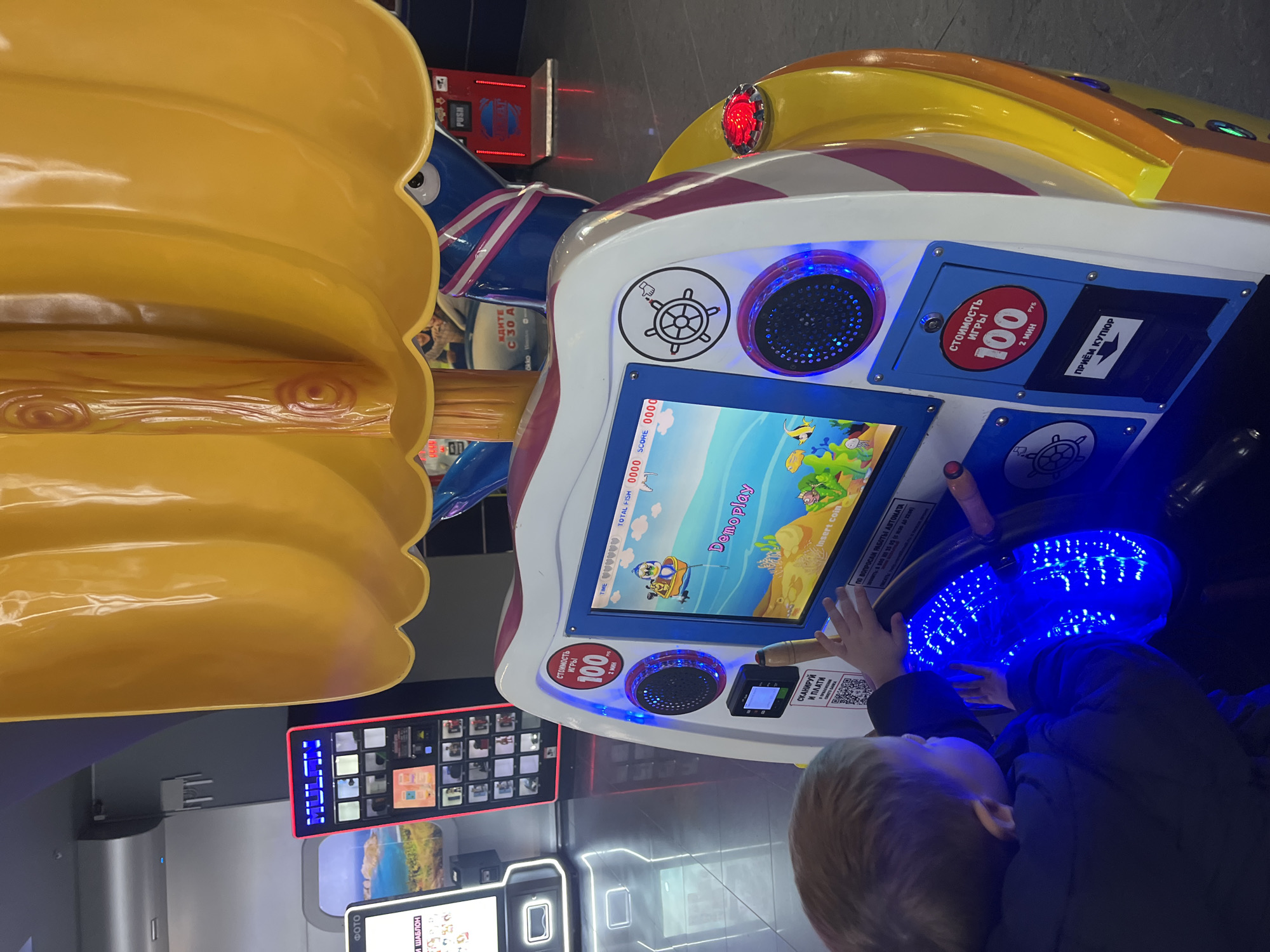 An indoor scene in what appears to be an entertainment center or arcade is captured, with the image rotated 90 degrees clockwise. The primary subject is a young child, seen from behind and the side, with light hair and wearing a dark jacket. The child's hands are placed on the illuminated steering wheel of an arcade game.
The arcade game is a kiddie ride simulator, designed with a boat or submarine theme. It features a bright yellow top section, a white main body with pink and purple stripes, and a blue base. The interactive console includes a screen displaying a colorful, animated underwater game with a character in a boat, various fish, a crab, and coral. The screen also shows "Demoplay" and prompts "Insert coin" and "TIME 000000 TOTAL FISH 0000 SCORE 0000." The steering wheel glows with bright blue lights, and blue-lit speakers are also visible on the console.
Adjacent to the screen are payment options. Text in Russian indicates "стоимость ИГРЫ 100 РУБ 2 мин" (Cost of game 100 RUB 2 min) and "ПРИЁМ КУПЮР" (Bill acceptor). Below the game screen, a QR code is visible with the instruction "СКАНИРУЙ И ПЛАТИ" (Scan and pay). Further text provides a contact for machine operation inquiries: "ПО ВОПРОСАМ РАБОТЫ АВТОМАТА" (For questions about the machine's operation), followed by a phone number and operating hours (05:00 до 22:00). A graphic depicting a hand turning a steering wheel is also present.
In the background, out of focus, other arcade machines and digital displays are visible. Text on these includes "ФОТО" (Photo), "И ШАБЛОН" (And template), "Okko" (a streaming service brand), "ЖДИТЕ С 30 А" (Wait from 30 A), and buttons labeled "PUSH" and "МЕН". The overall setting suggests an evening or indoor environment with artificial lighting. The presence of Russian text aligns with the given location context of Ulyanovsk, Russia.