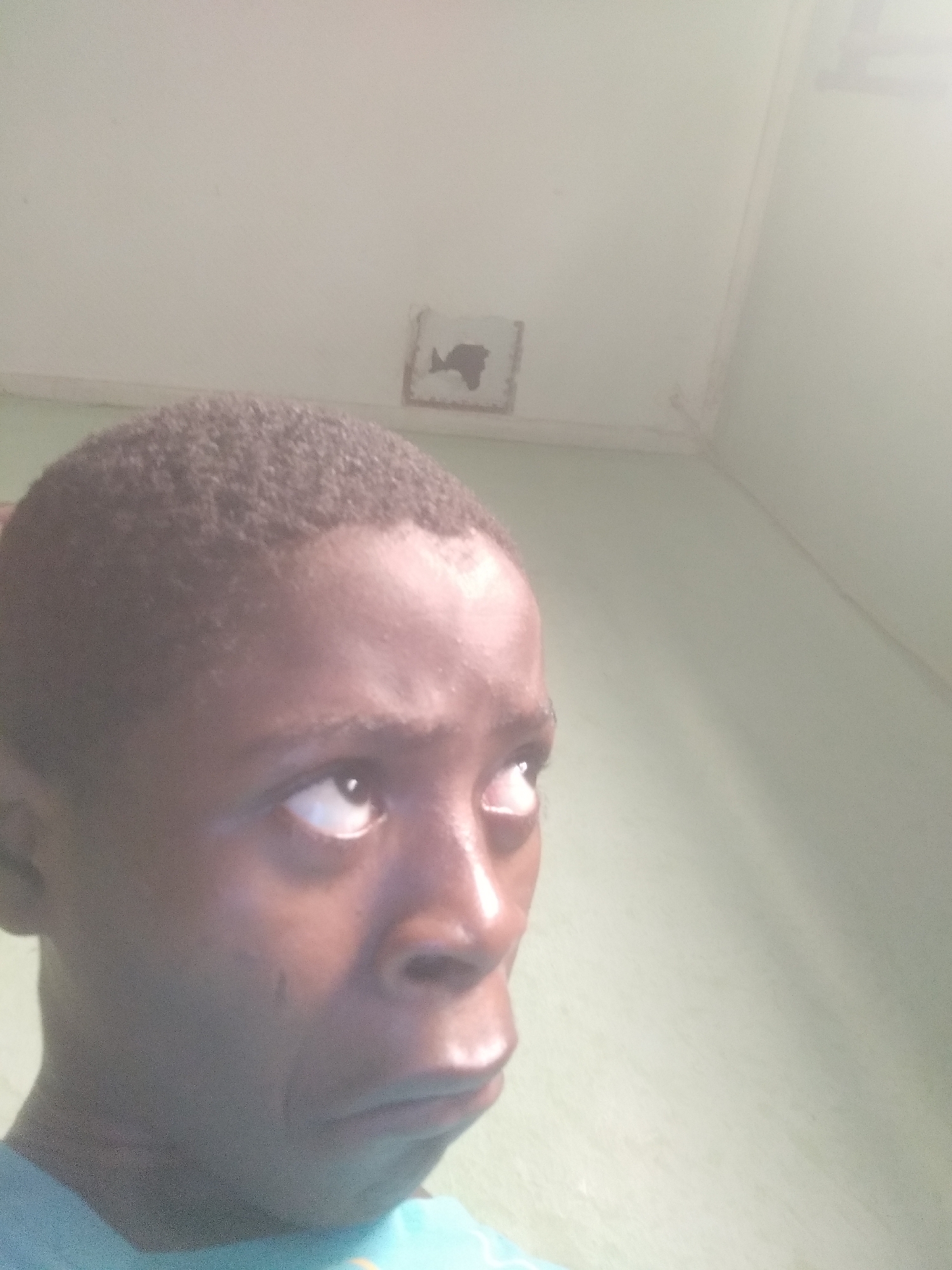A close-up, slightly blurry portrait of a young Black man with short, dark hair. He's looking upwards and to the right, with his eyes slightly wide. His expression appears contemplative or slightly doubtful. The background consists of a white ceiling and a pale green wall. A discolored square is visible on the ceiling. The image does not confirm a location in Shangisha, Nigeria.