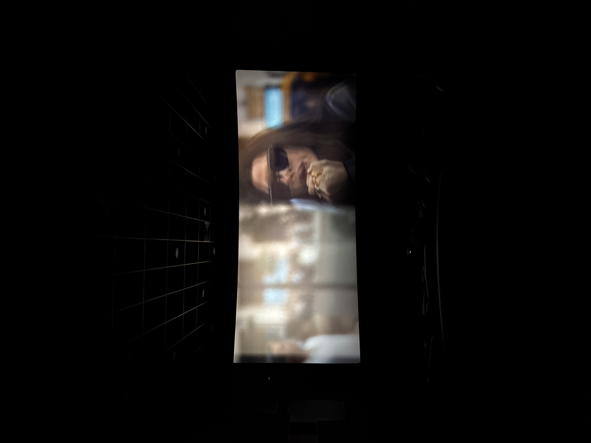 A person with long hair and sunglasses is visible on a screen in a dark room. The person is wearing rings on their fingers. The screen is vertically oriented. A gridded wall is on the left. A portion of seating is visible at the bottom of the frame. The city and country are not discernible in this image.