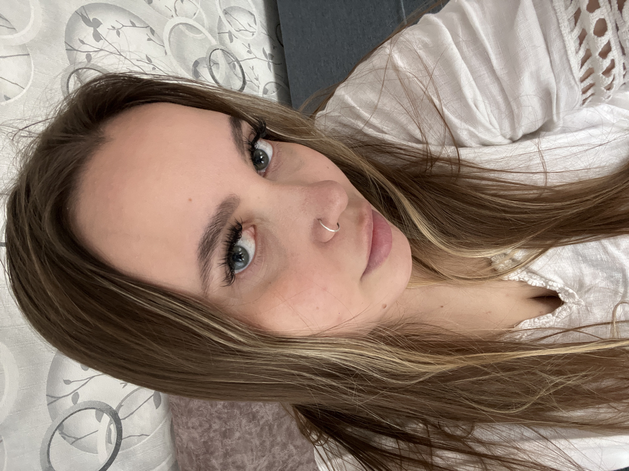 This close-up portrait captures a young woman in an indoor setting, likely in Port-Katon, Russia. She is positioned from the chest up, lying down with her head tilted, and engaging directly with the camera. Her expression is calm and composed.
The woman has long, flowing light brown hair with subtle blonde highlights that frames her face and spreads around her. Her prominent features include striking light blue or grey eyes, enhanced by long, dark eyelashes and neatly shaped eyebrows. A silver hoop nose ring adorns her left nostril, and her lips are softly pink and slightly parted. She is wearing a light-colored, possibly white, top with delicate texture or lace detailing visible on the right side.
The background features a patterned fabric, suggesting a bedsheet or pillowcase, with a decorative motif of grey branches, leaves, and bird silhouettes on a lighter base. A darker, solid-colored surface is also visible in the upper right background. The lighting is soft and even, contributing to a relaxed and intimate atmosphere. There is no visible text in the image.