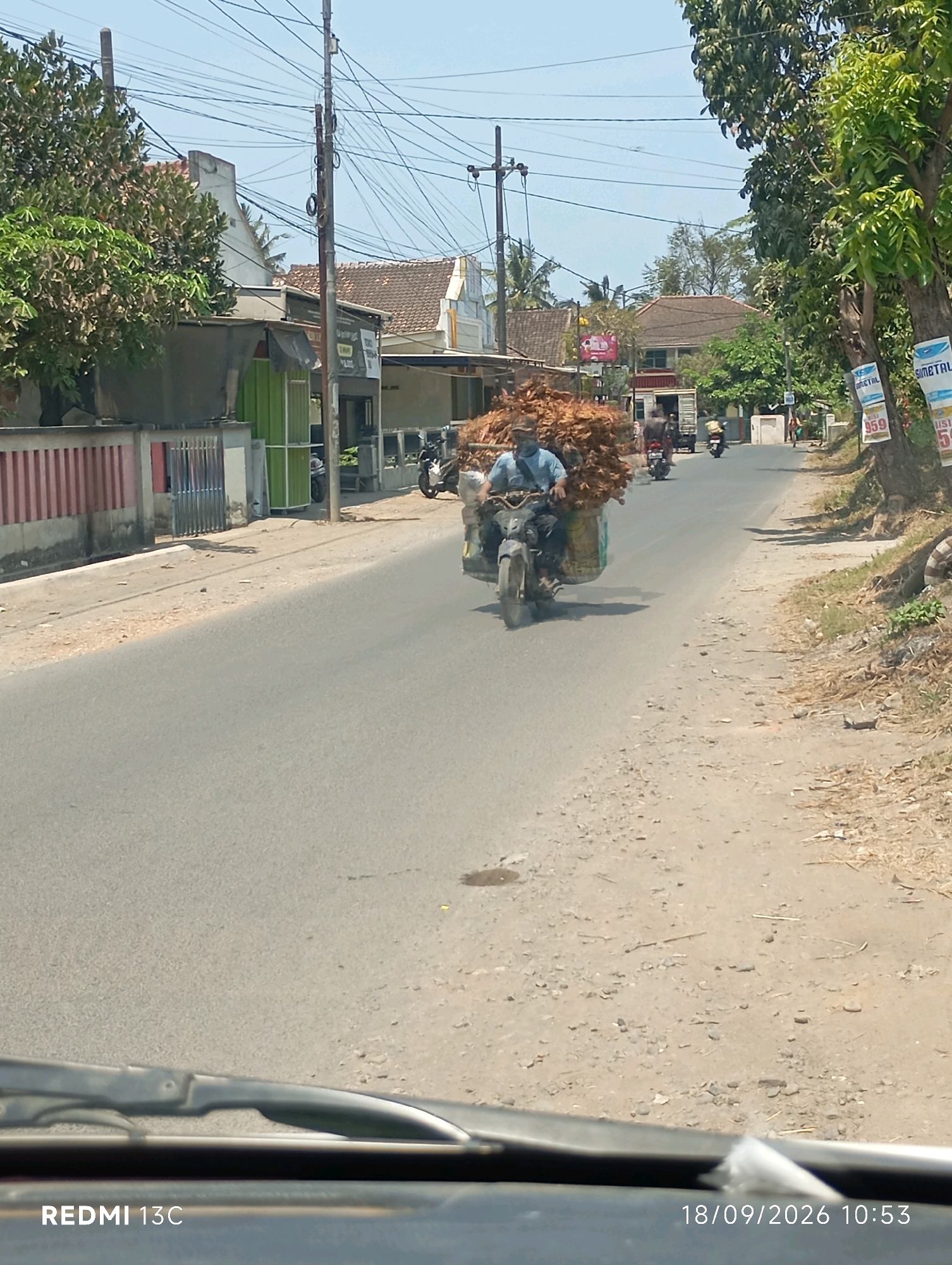 A motorcycle with a large load of dried leaves is being ridden down a paved road in Sumberrejo, Indonesia, on September 18, 2026, at 10:53 AM. The rider, wearing a blue shirt and a mask, is seated on the motorcycle, which is equipped with a sidecar or modified passenger seat to hold the substantial cargo. The load consists of tightly packed, dry, brown leaves or similar plant material.
The road is flanked by a dusty shoulder and vegetation on the right, with buildings and trees on the left. Power lines crisscross the sky above. A few other vehicles, including a truck and other motorcycles, are visible further down the road. The weather appears to be clear and sunny. The image is taken from a low angle, possibly from inside another vehicle, as indicated by the presence of a windshield wiper and a portion of a dashboard in the foreground.