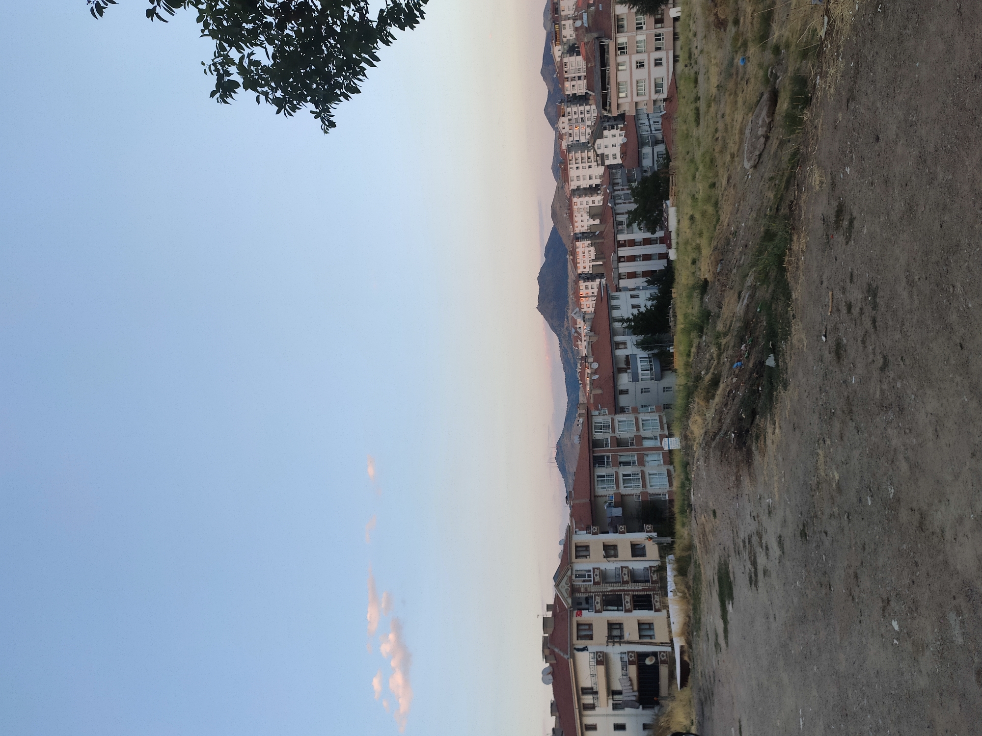 The image depicts a residential area in Mamak, Turkey, under a clear, pale blue sky with a few wispy pink clouds near the horizon. In the foreground, a dirt path and uneven, dry grassy terrain suggest an undeveloped or transitional space. Beyond this, a row of multi-story apartment buildings, typical of urban Turkish architecture, are visible. These buildings feature reddish-brown tiled roofs, white and beige facades, and numerous balconies. Some buildings have satellite dishes on their roofs, and clothes hang from some balconies, indicating occupancy. In the background, rolling hills and mountains form the city's natural backdrop. A tree with dark green leaves is visible in the upper right corner of the frame, its branches silhouetted against the sky. The scene appears to be captured during late afternoon or early evening, given the soft lighting and the colors in the sky. There are no people visible in the image.