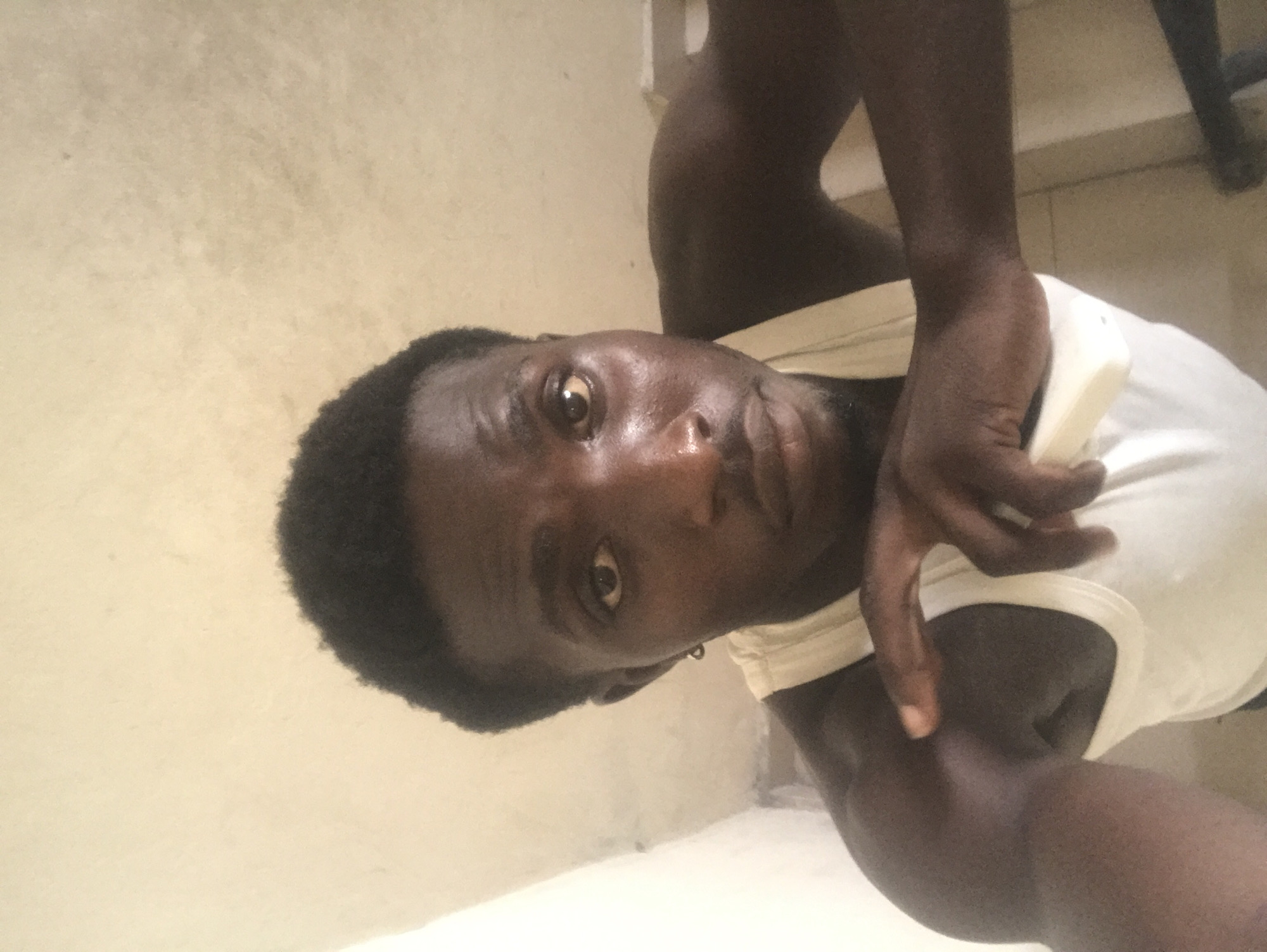 A dark-skinned man wearing a white tank top is captured in a close-up, head-and-shoulders selfie. He has short, dark, curly hair and a faint beard. His gaze is directed towards the camera, with his right hand resting on his chest. The background is a plain, light-colored wall, suggesting an indoor setting. The lighting is somewhat dim, casting shadows on his face. There is a hint of a white wall to his left. The man is wearing a small hoop earring in his left ear.