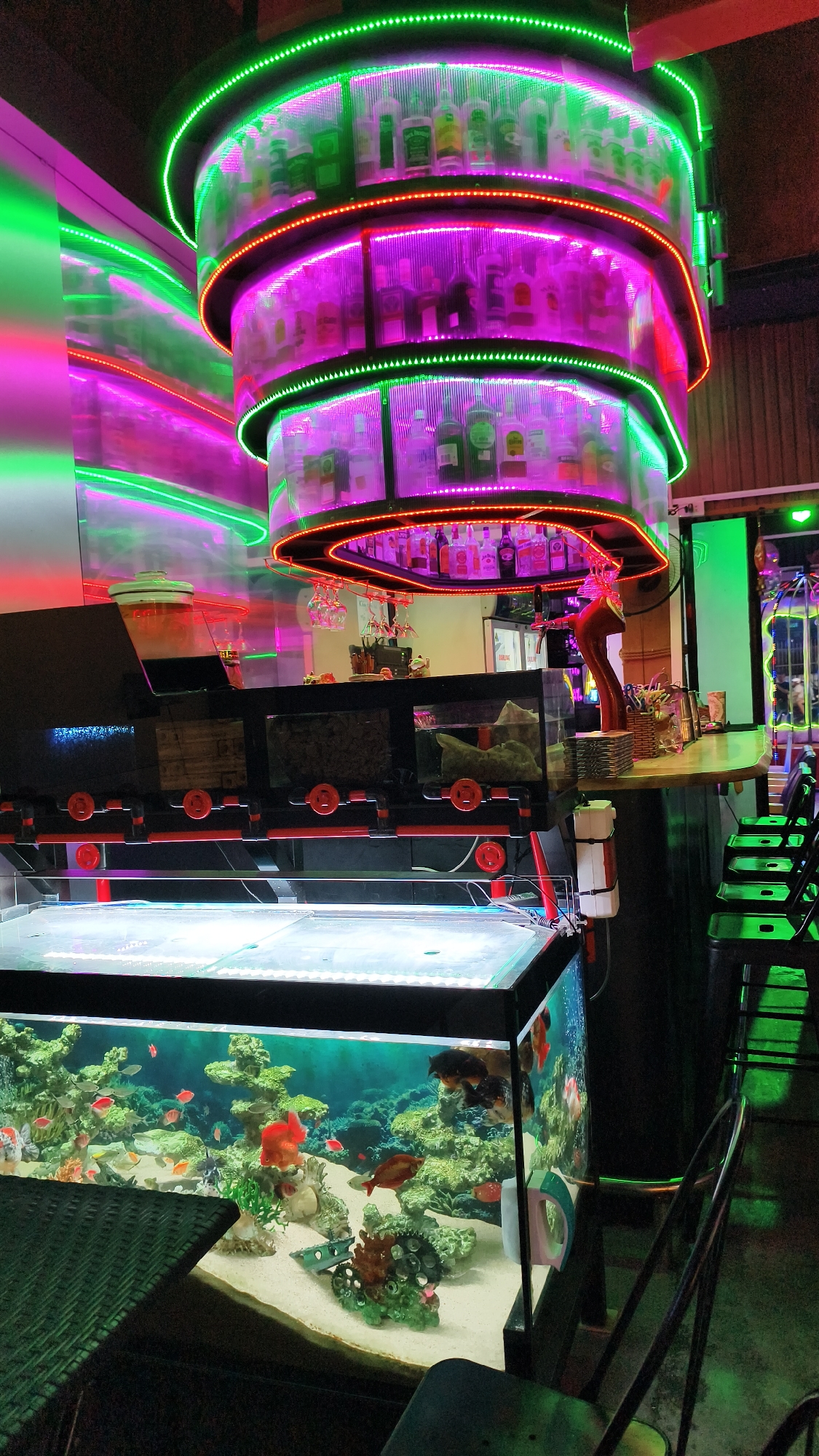 The image depicts the interior of a bar or restaurant, likely located in Nha Trang Ward, Vietnam, as indicated by the metadata. The scene is characterized by vibrant neon lighting, primarily in green and purple hues, which illuminate a multi-tiered bar display stocked with bottles of liquor. Below the bar, a large aquarium is prominently featured, filled with various colorful fish and aquatic plants, creating a lively underwater environment.
The bar area itself has a polished countertop and several stools arranged in front of it. A beer tap with a red handle is visible on the bar. To the left of the bar display, a reflective surface with green and pink light streaks suggests another illuminated architectural feature or wall decoration.
In the foreground, a portion of a dark, textured table is visible, likely a seating area for patrons. The floor appears to be dark and possibly concrete or tile. The overall ambiance is modern and energetic, with the aquarium adding a unique natural element to the establishment. No people are visible in the image.