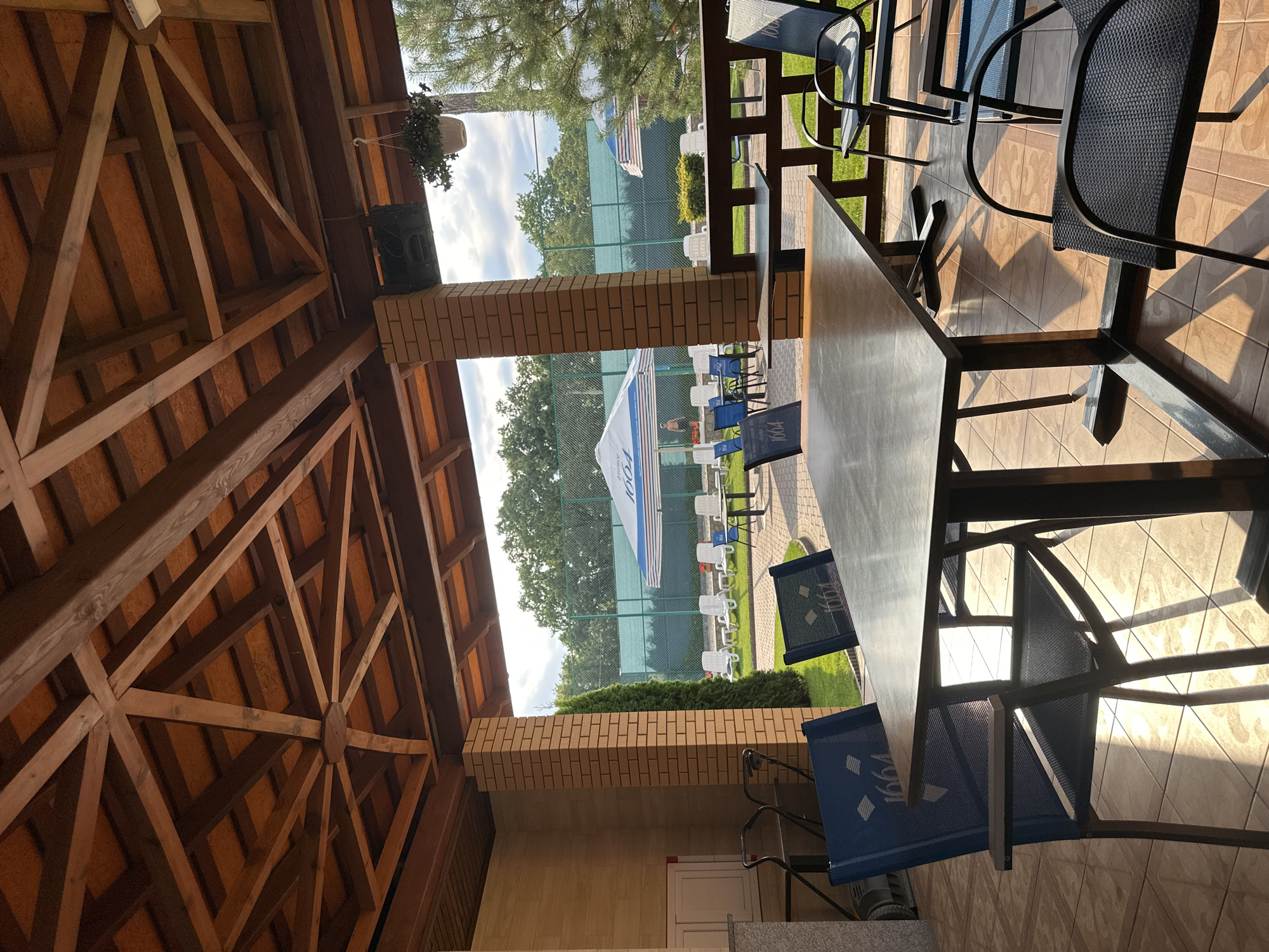 The image depicts an outdoor seating area under a wooden pergola, likely at a resort or cafe, with a tennis court visible in the background. The scene is set in Khmilnyk, Ukraine. In the foreground, a dark rectangular table is surrounded by chairs. Two prominent blue chairs with a "1664" logo are visible on the left, with another black mesh chair to the right. The sunlight casts long shadows across the tiled floor, indicating it is late afternoon or early morning.
Beyond the seating area, lounge chairs are arranged on a manicured lawn, some under large white and blue striped umbrellas. A person, possibly a man, is standing near one of the umbrellas in the middle ground. A tennis court fence runs along the background, with trees and a cloudy sky visible beyond it. To the left of the seating area, a brick pillar supports the pergola, and a light-colored wall with a cabinet is visible. A trolley with wheels is parked to the far left. The overall atmosphere suggests a relaxing outdoor space.
