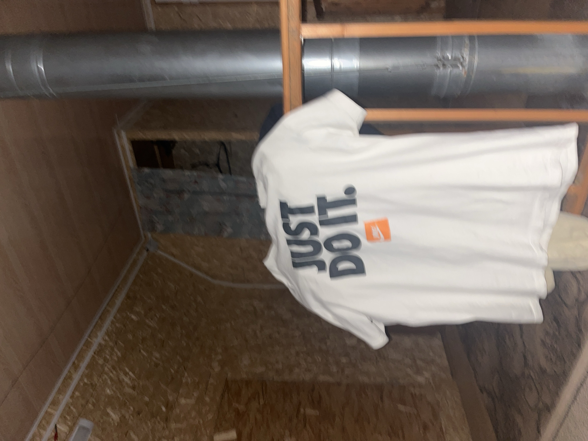 The image shows a white t-shirt with the "JUST DO IT." slogan in large, dark letters and a small orange Nike logo. The t-shirt is hanging on a wooden railing, with a large, silver metal pipe visible behind it. The background consists of wood-paneled walls and a wooden ceiling, suggesting an interior space such as a room under construction or a workshop. The overall lighting is dim. This scene is not located in Chaikovsky, Russia, as there is no specific information or context in the image to suggest this location.