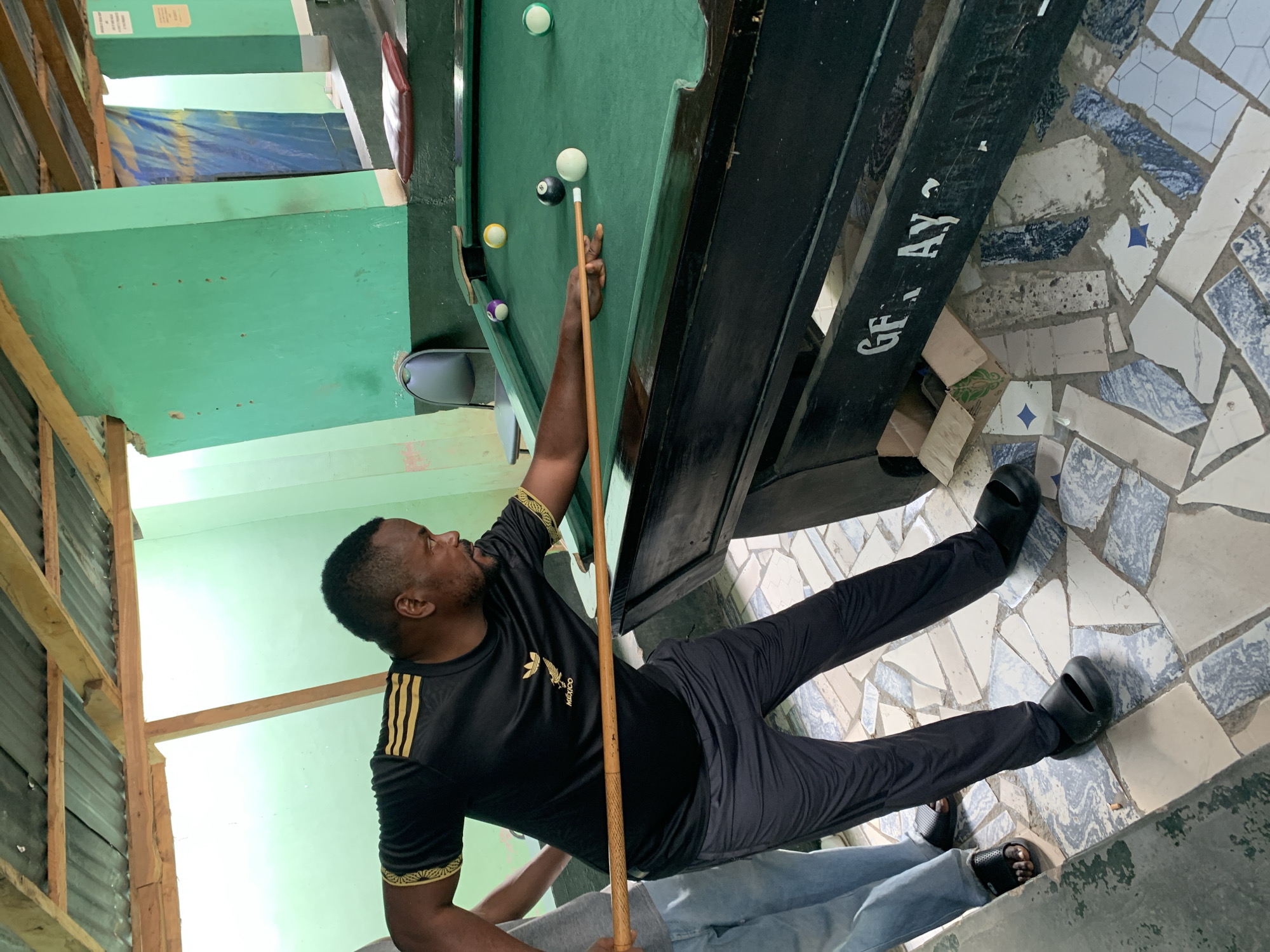 A man wearing a black shirt and pants is playing pool in a room in New Stadium, Nigeria. The pool table has several balls on it. The floor is covered in irregular tiles. Another person is standing at the edge of the frame.