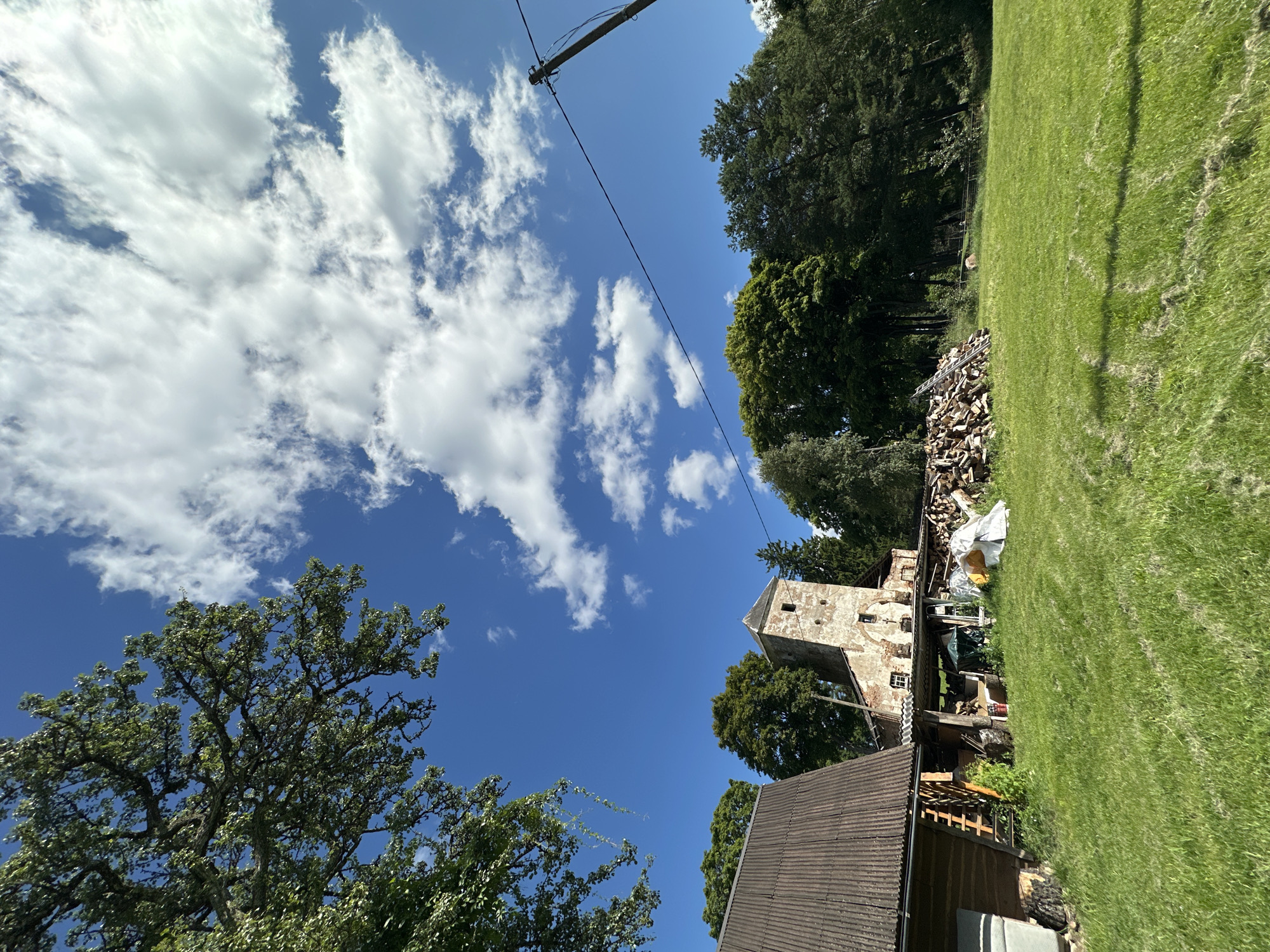 In Norviliškės, Lithuania, a wide-angle shot captures a rural scene under a bright blue sky dotted with fluffy white clouds. In the foreground, a lush green lawn slopes gently upwards. To the left, a rustic wooden building with a corrugated metal roof stands partially in view. Adjacent to it, an old stone tower, possibly part of a historic structure, rises prominently, showing signs of age and weathering. A large pile of cut firewood is stacked near the tower, with some tarpaulins and other materials scattered around. A power line stretches across the frame, supported by a wooden pole on the right side. The background is dominated by a dense forest of green trees. The overall impression is one of a serene, somewhat unkempt, rural landscape.