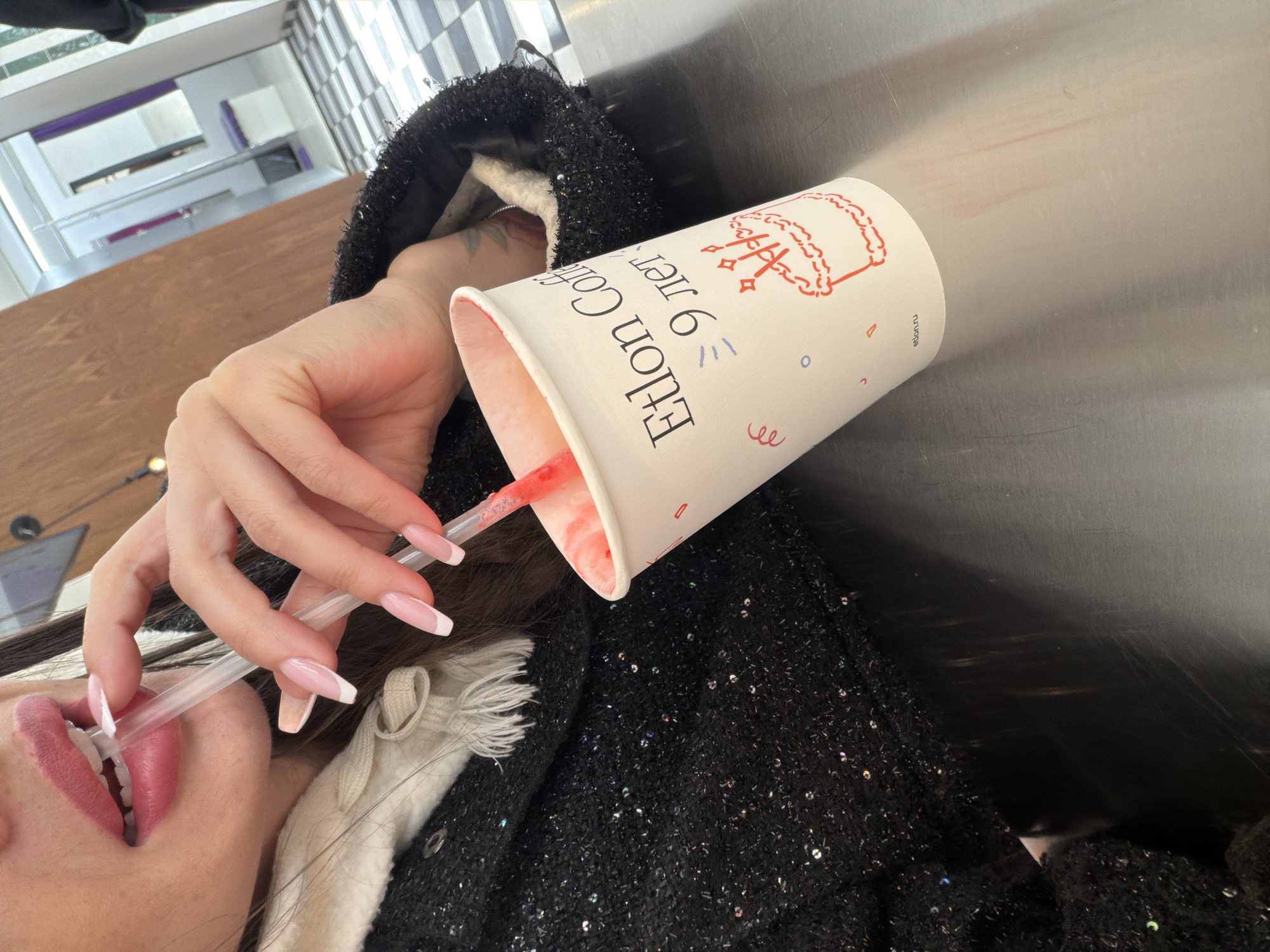 A person is drinking from a white paper cup with a red straw. The cup is labeled "Eilon Coffee 9 лет", indicating that the location is in the city of Saint Petersburg, Russia. The person's hand is holding the cup and straw. The person has long, manicured nails with a white tip design. The person is wearing a black jacket or coat with sequins. The background is blurry and shows parts of a building interior.