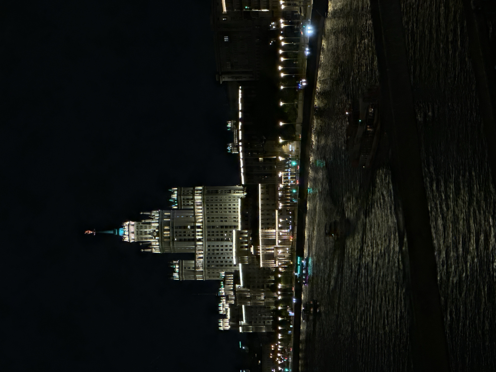 The image captures a night scene along the Moscow River in Russia. Prominent buildings line the riverbank, illuminated with numerous lights, reflecting on the water. The Kotelnicheskaya Embankment Building, a Stalinist skyscraper, is visible. The river itself appears dark and textured, suggesting some movement or ripples on the surface. Streetlights and other architectural lighting contribute to the bright line that separates the dark sky from the river. The overall impression is one of a vibrant, urban environment at night.