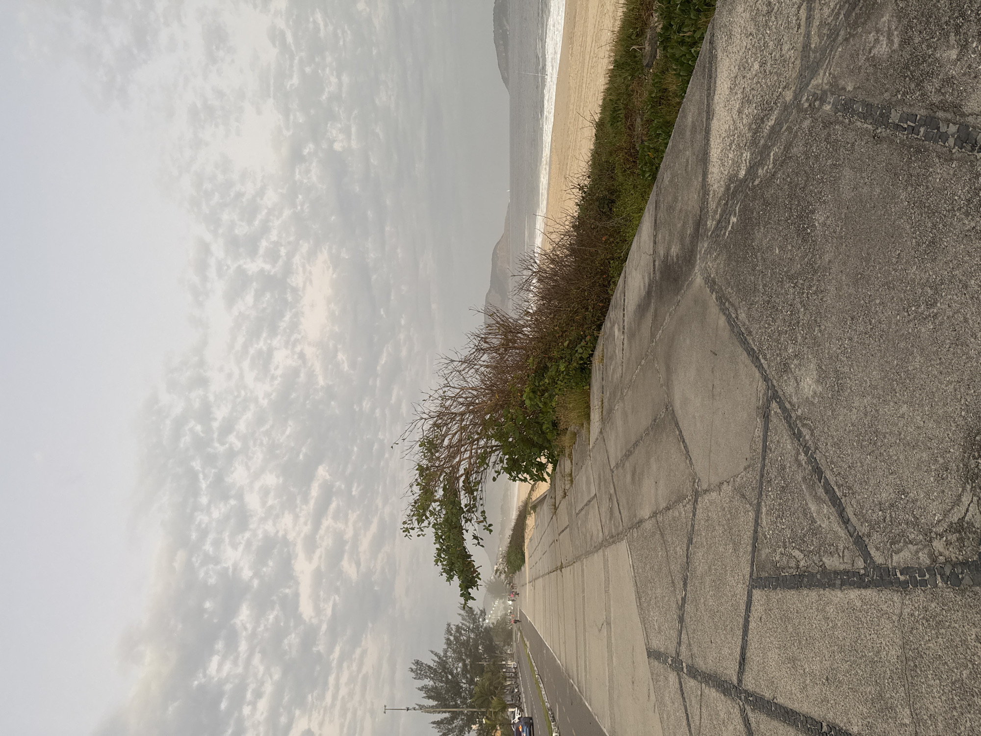 This image depicts a cloudy day at Icaraí Beach in Niterói, Brazil. A paved promenade runs alongside the beach, with a road and trees on the other side. The beach itself is sandy, with waves from the ocean lapping at the shore. In the distance, hills and islands are visible. Several people can be seen walking and cycling on the promenade, and a few cars are driving on the road. The overall atmosphere is calm and serene.