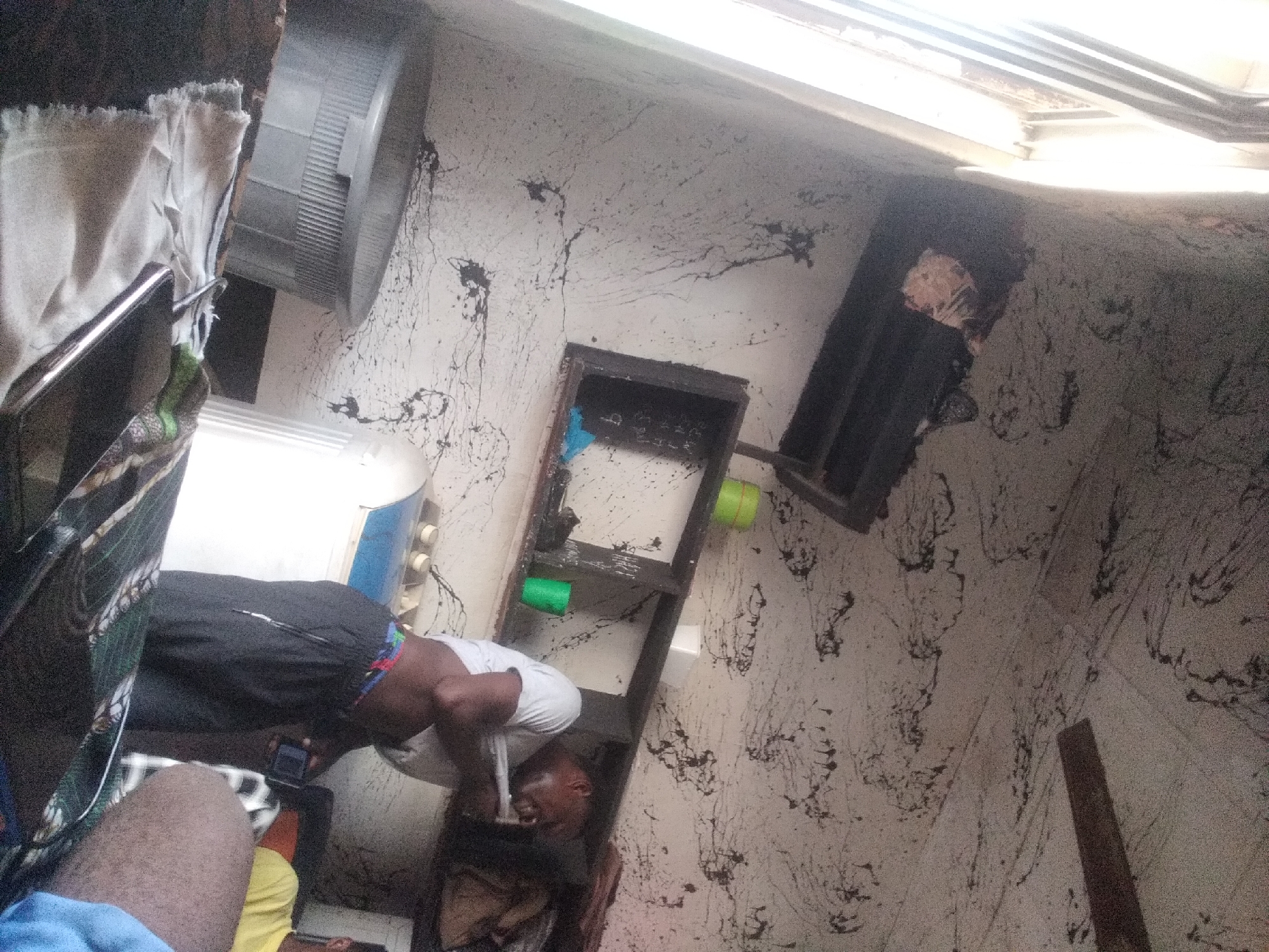 In Jalingo, Nigeria, an indoor scene depicts a cluttered, possibly dilapidated environment. A person is lying down, partially obscured by clothing and furniture. They are wearing black shorts with a colorful waistband and a white t-shirt. Their head is resting on something dark, and their facial expression suggests they are speaking or reacting to something. Their right hand is holding a small black electronic device, possibly a phone.
The walls are textured and appear stained with dark, irregular splotches, giving the impression of decay or neglect. A metal ventilation fan is visible in the upper left corner. There is a shelving unit with dark wooden frames and what appear to be some plastic containers, one green and one light blue, placed on it. Light streams in from above, suggesting it might be daytime. The overall atmosphere of the scene appears unkempt and somewhat somber. The context of Jalingo, Nigeria, places the scene within a specific geographical region, though no explicit visual cues within the image directly confirm this location. No visible text is present in the image.