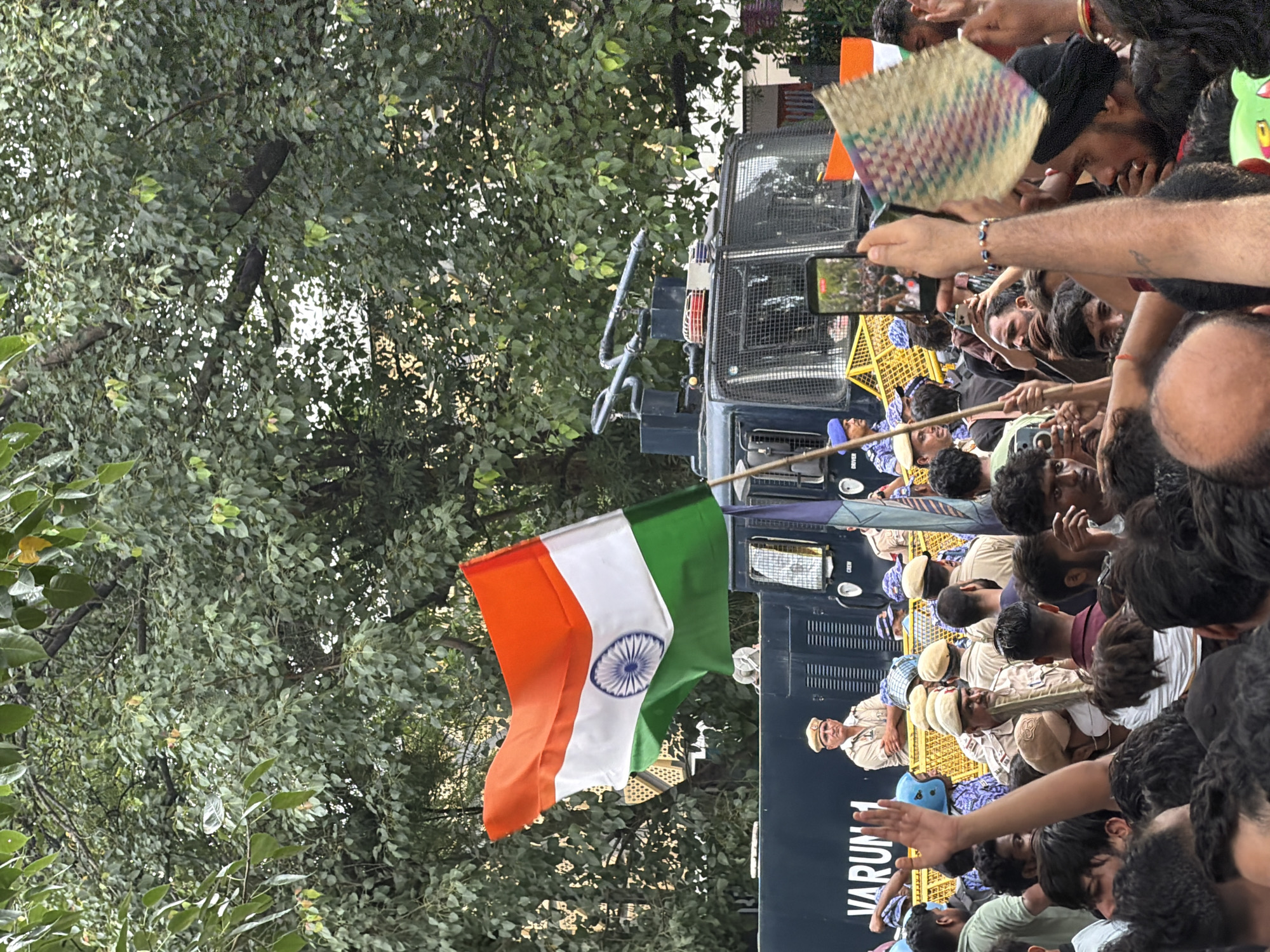 The image depicts a crowd of people gathered in an outdoor setting. A large, dark-colored vehicle, possibly a police or water cannon truck, is visible in the background. An Indian flag is prominently displayed, waving in the foreground. Several individuals in uniform, likely police officers, are interspersed among the crowd and near the vehicle. Some people are holding mobile phones, suggesting they are capturing the event. The overall atmosphere appears to be that of a public gathering or protest. The presence of the Indian flag and police presence suggests a location in India, likely a city. The text "VARUNAL" is visible on the side of the dark vehicle, which could be a company name or model. The setting is outdoors with trees and foliage in the background.