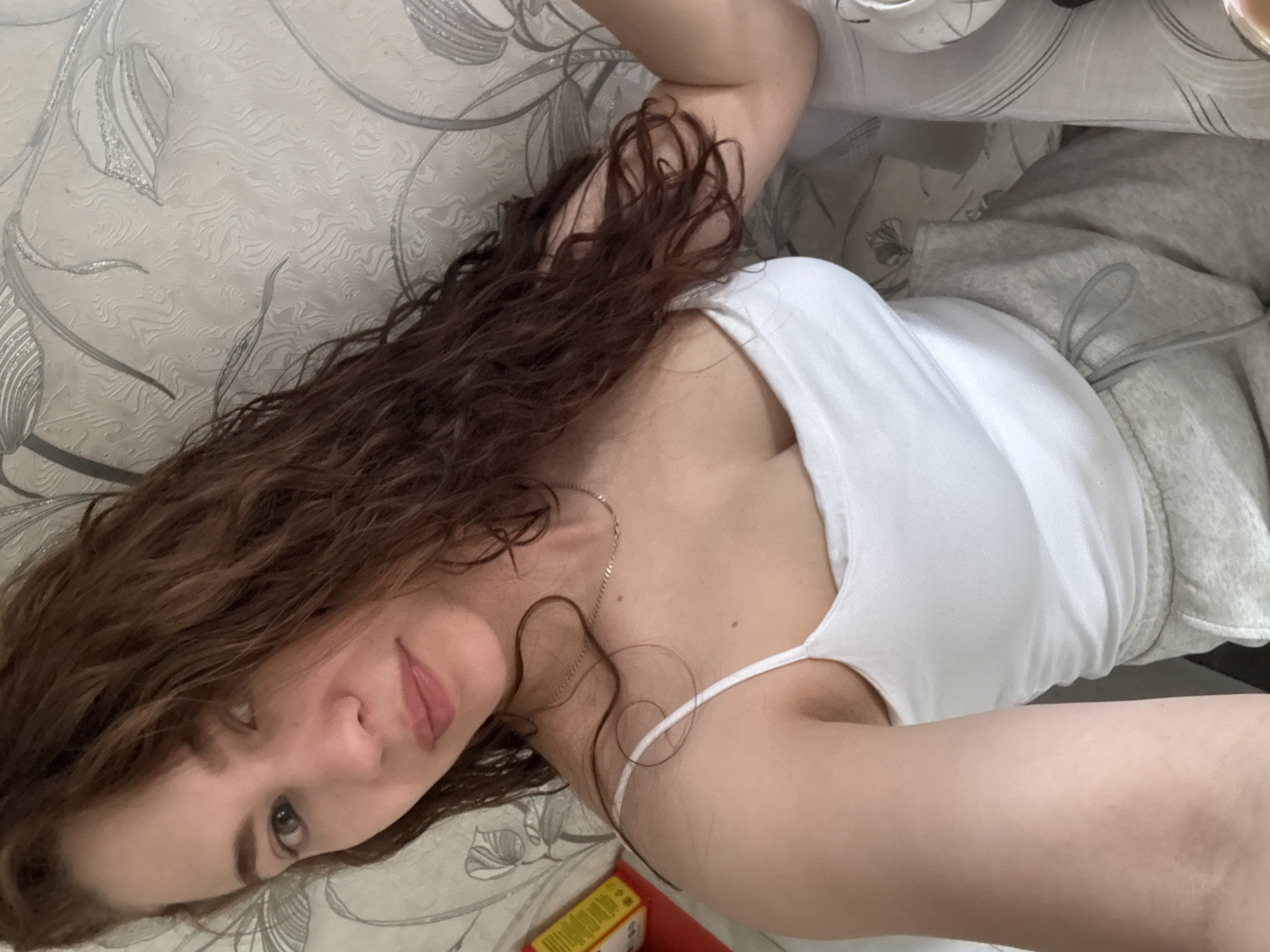A woman lies on a patterned surface, presumably a bed or sofa, wearing a white tank top and gray sweatpants. She has long, curly brown hair. A portion of a yellow and red box is visible in the lower left corner of the frame. The background appears to be a patterned wallpaper. The location is not discernible as Nizhnyaya Gar', Russia from the given image.
