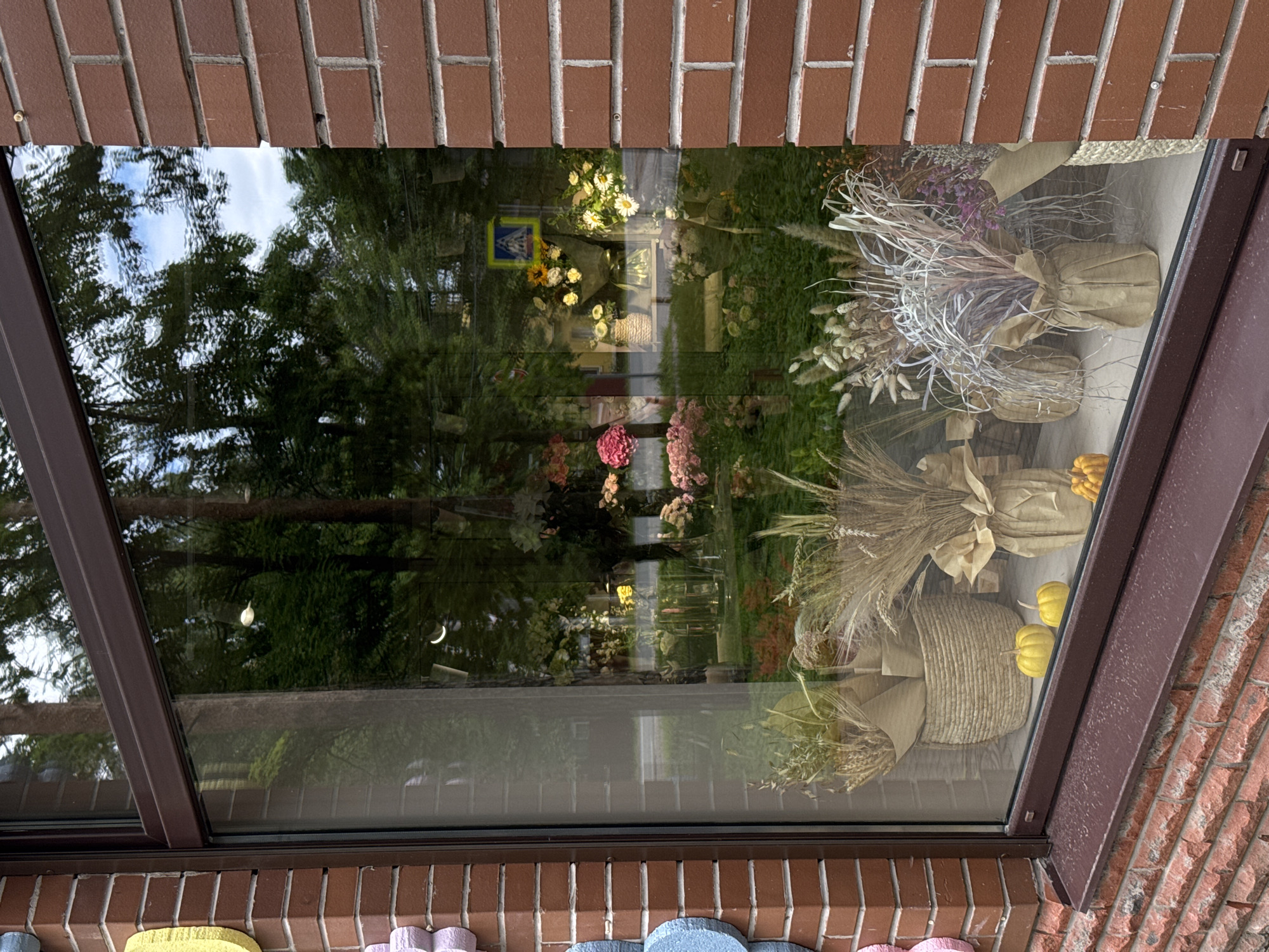 This is a medium shot, eye-level exterior photograph of a storefront window with reflections of trees and sky. The window is framed by reddish-brown brick. To the left of the window, decorative elements in pastel blue, pink, and yellow are attached to the brick wall. The window displays dried floral arrangements in woven baskets and brown paper-wrapped vases, along with two small yellow pumpkins on the windowsill. Inside the store, visible through the window, are more floral displays, including pink and yellow flowers in vases and on shelves. A pedestrian crossing sign is visible in the reflection in the upper right portion of the window. The scene appears to be set during the daytime.