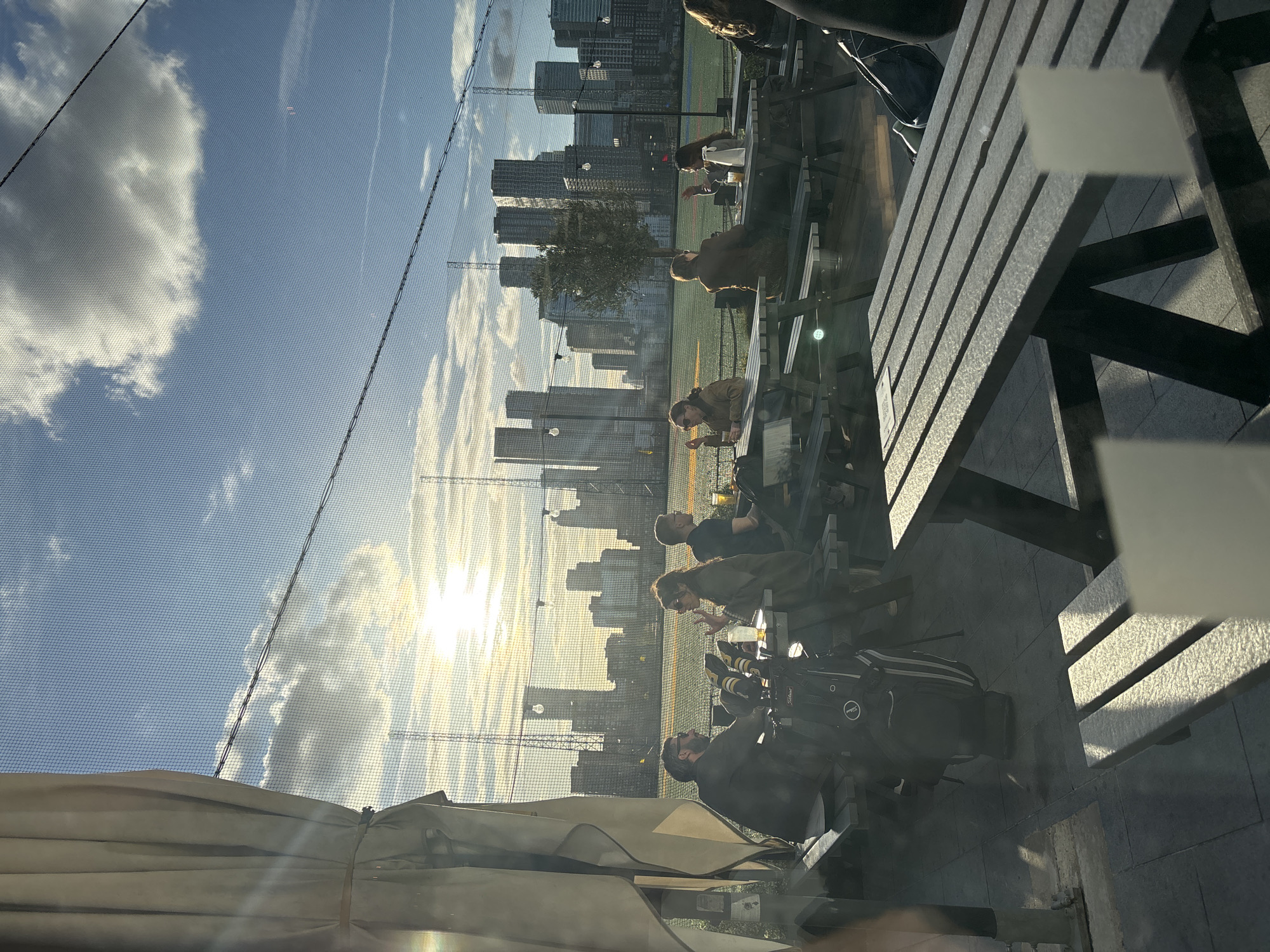 The image captures an outdoor scene at what appears to be a golf driving range, with a city skyline in the background, likely in London, UK. The shot is taken from a slightly elevated perspective, looking across a seating area towards the distant city.
In the foreground, several people are seated at wooden picnic-style tables and benches, some under the shade of light-colored awnings or umbrellas. They appear to be socializing or relaxing. One person is visible on the far right, partially obscured by reflections. Near the center, a black golf bag with the "Titleist" logo is placed beside one of the tables, suggesting that the location is either a golf facility or a venue popular with golfers.
A large, fine-mesh net, typical of a golf driving range, stretches across the mid-ground, separating the seating area from a green field. Beyond this field, a dense cluster of modern skyscrapers forms a prominent city skyline, silhouetted against the bright sky. The sun is low in the sky, appearing very bright and creating a strong lens flare from the right side of the frame. This intense sunlight indicates either late afternoon or early evening, casting long shadows across the seating area and illuminating the clouds in the blue sky. Some scattered clouds are visible, with prominent light reflections on their undersides.
The overall atmosphere is one of leisure and relaxation in an urban recreational setting.