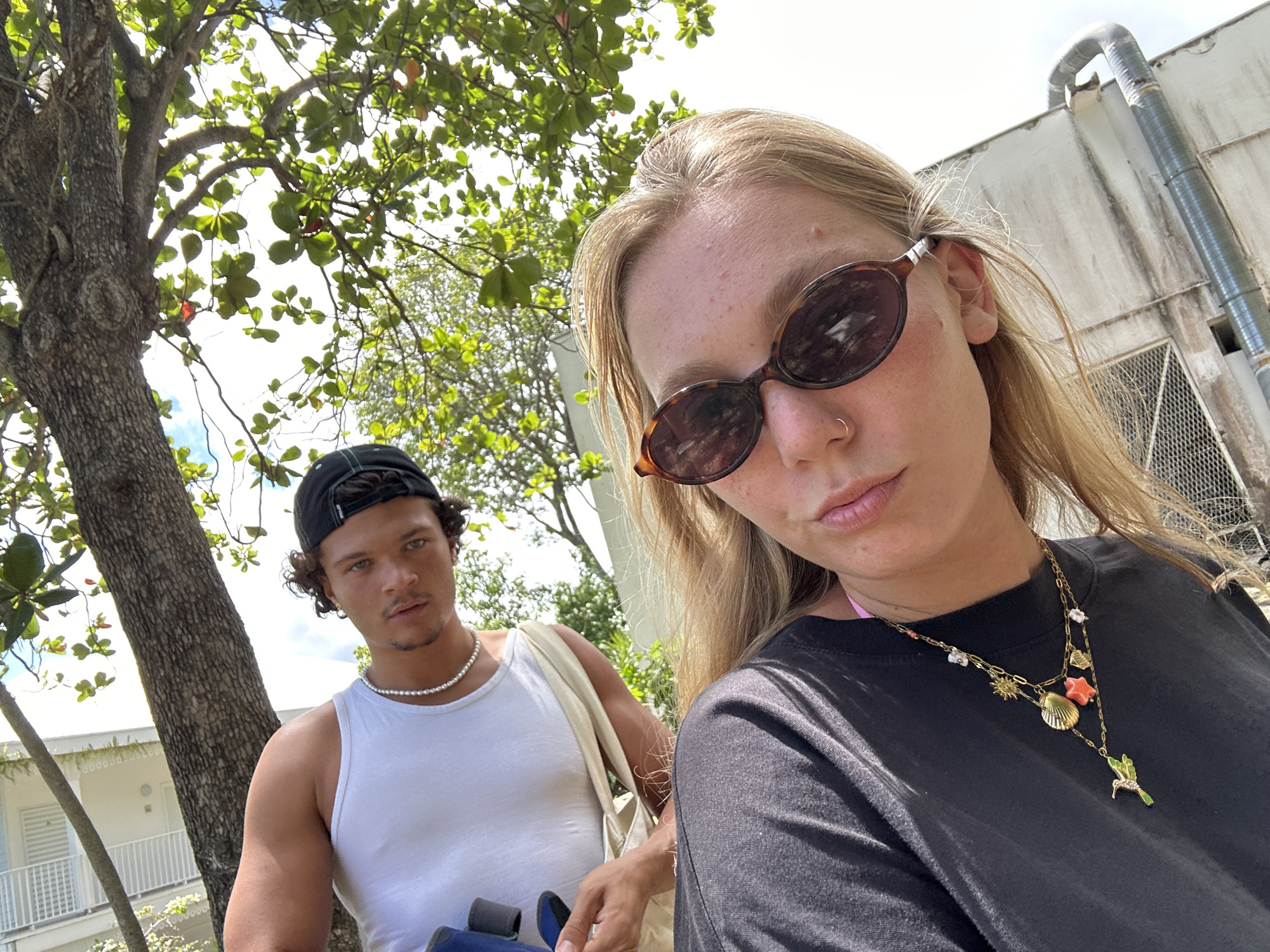 Two individuals are captured in a selfie-style photograph. The person in the foreground, presumably the one taking the photo, is fair-skinned with light blonde hair and wears dark, oval sunglasses. A black t-shirt and a gold necklace adorned with multiple pendants are visible. Behind them, another person with darker complexion, dark curly hair, and a black baseball cap is present. They wear a white tank top and a pearl necklace. A beige tote bag hangs from their shoulder.
A tree with green foliage is situated to the left of the frame, while a light-colored building with a vertical metal pipe running along its exterior is visible in the background on the right. The photograph is taken outdoors in daylight. While the specific location within Sainte-Anne, France, is not discernible from the image, the presence of trees and buildings suggests an urban or suburban environment.