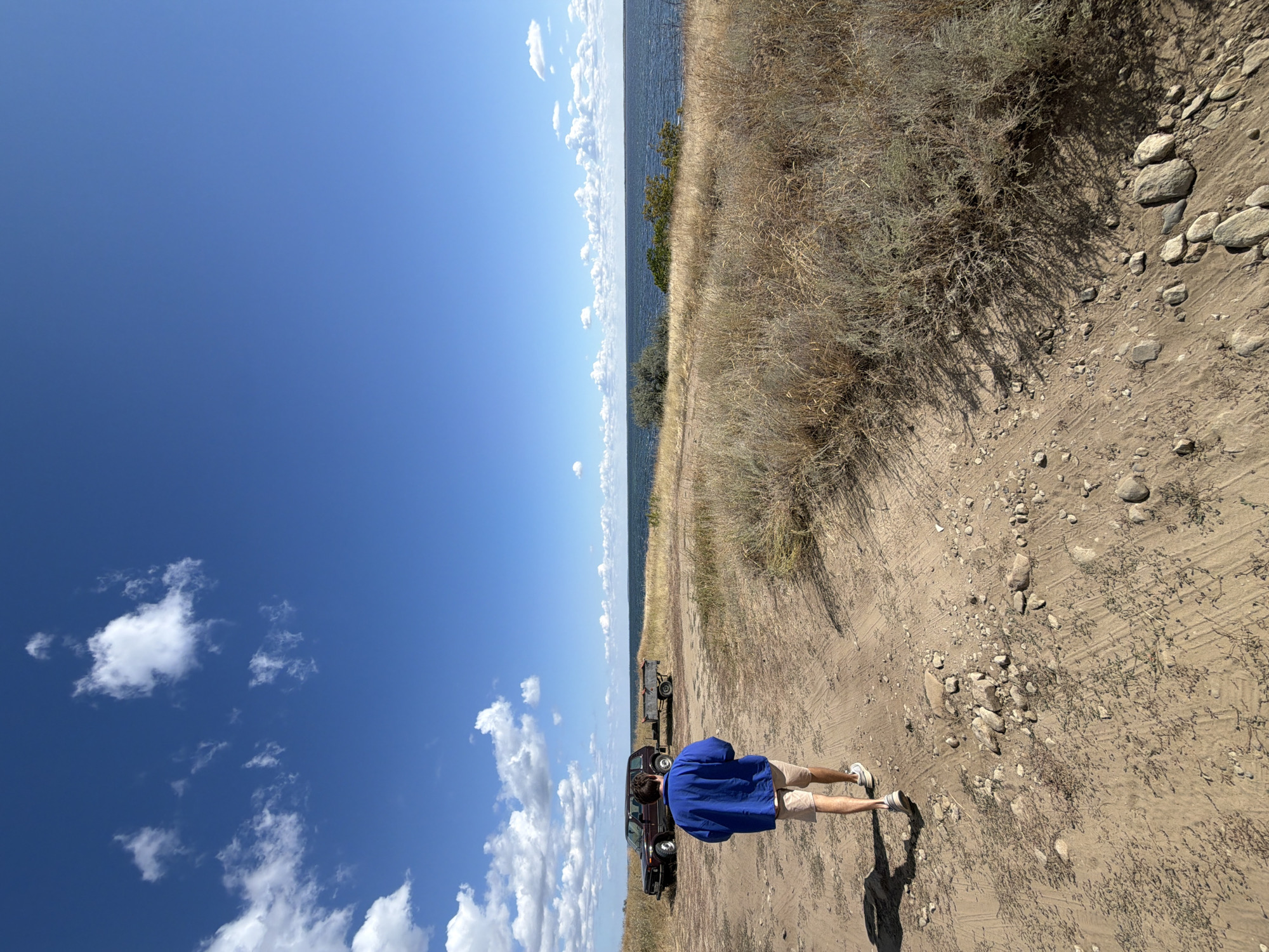 A young man wearing a blue jacket and tan shorts walks away from the camera on a dirt path. He is heading towards a body of water, possibly a river or sea, under a bright blue sky with scattered white clouds. In the background, a dark-colored SUV and a trailer are parked near a grassy embankment. The terrain is dry and covered with sparse vegetation and rocks. The scene appears to be a natural, undeveloped area, possibly on the outskirts of a city. The location is identified as Kamyshin, Russia.