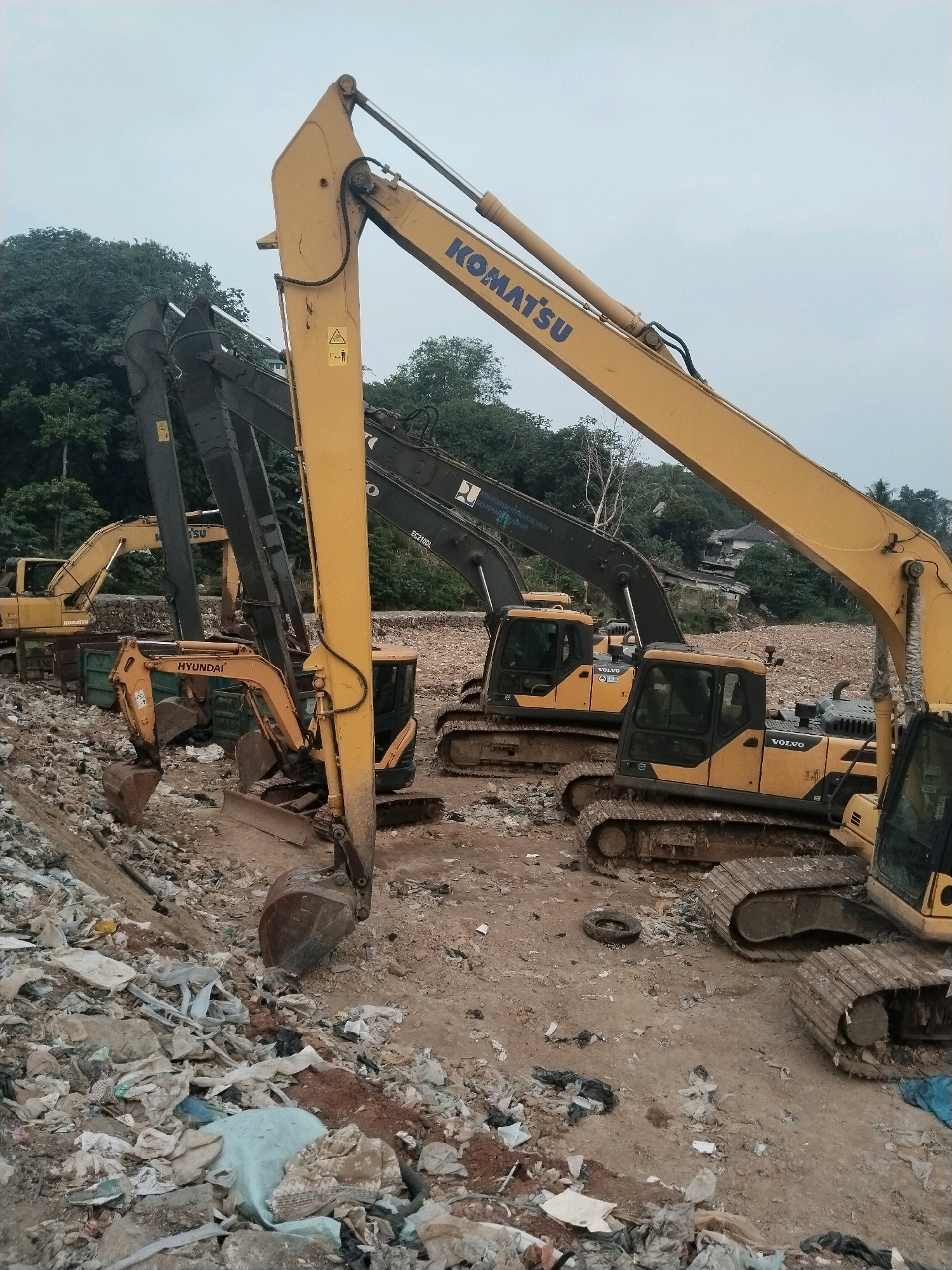 The image shows a landfill site in the city of South Tangerang, Indonesia. Multiple excavators, including Komatsu and Volvo models, are present, some actively digging and moving debris. The ground is covered in a mixture of dirt, rubble, and various types of waste, including plastic bags, fabric, and construction debris. Trees and houses are visible in the background, suggesting the landfill is located on the outskirts of a residential area. The sky is overcast, indicating potentially humid or rainy weather. There are no people visible in the image.