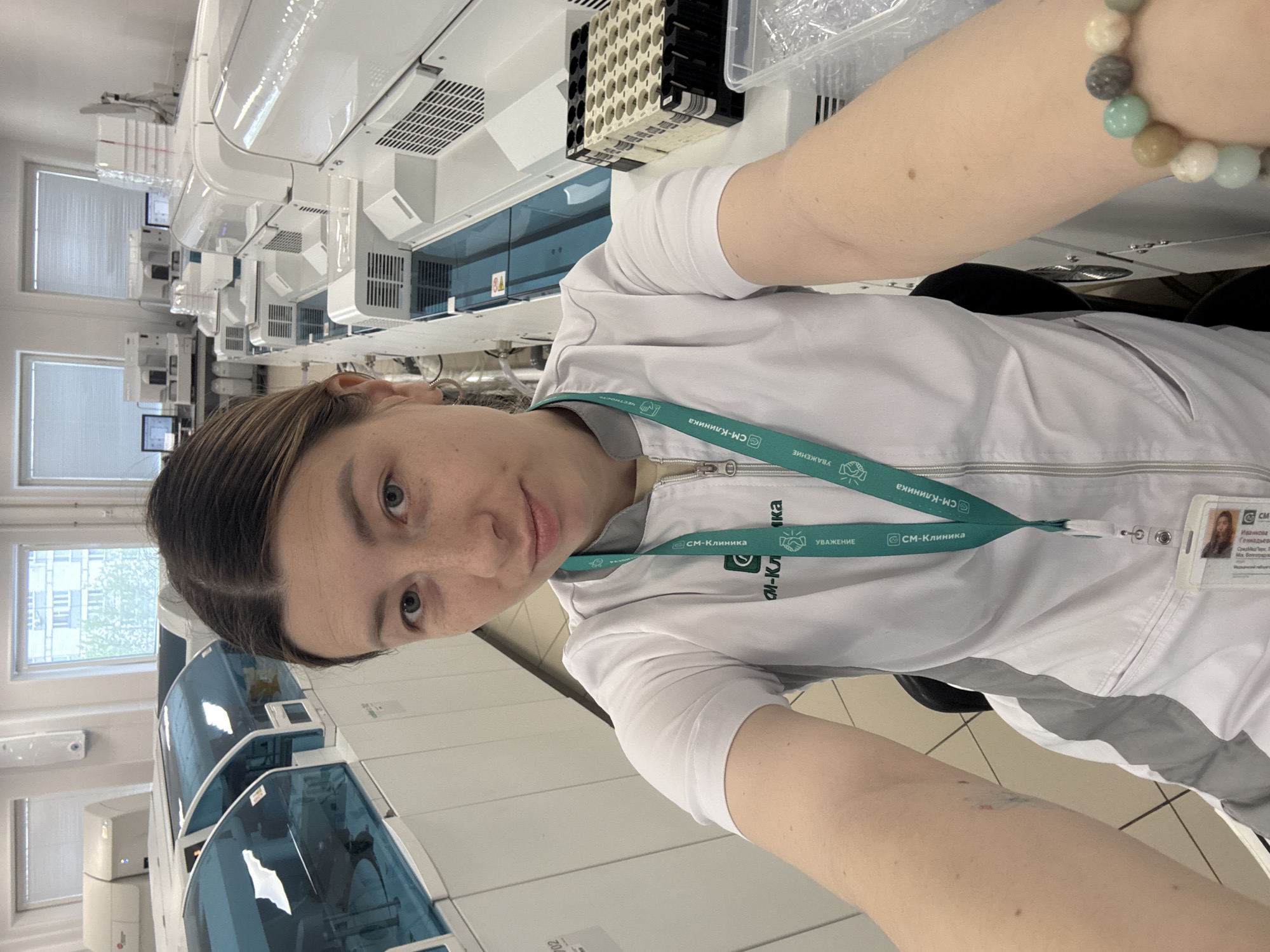 A woman wearing a white medical coat with a "CM-Clinica" lanyard is taking a selfie in what appears to be a medical laboratory. The background shows rows of laboratory equipment with windows in the distance. The "CM-Clinica" emblem on her lanyard and coat suggests that this image was taken in a medical facility located in the city of Moscow, Russia, where "CM-Clinica" has branches.