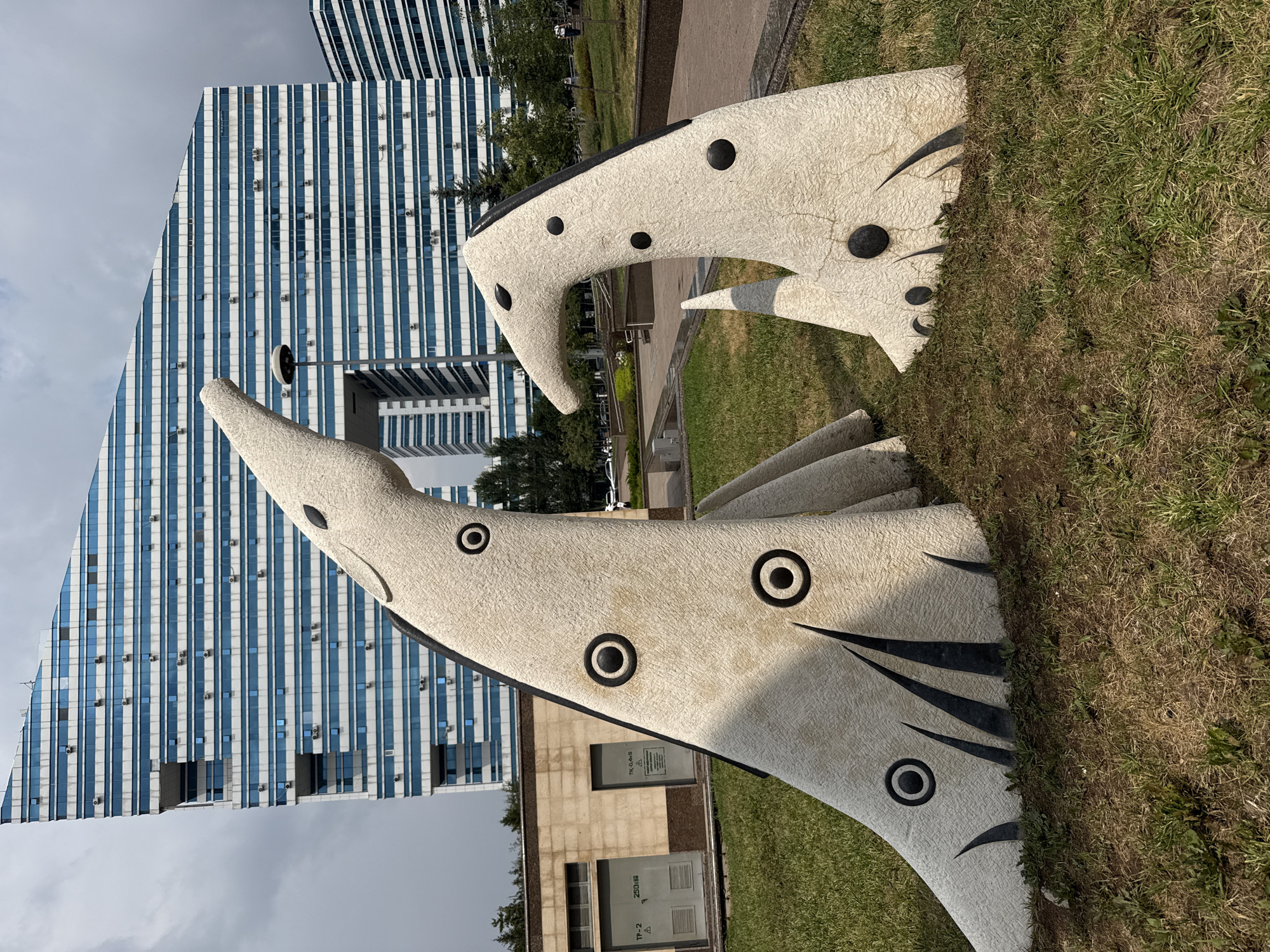 This is a medium shot, eye-level, outdoor photograph of two abstract sculptures made of a light-colored, textured material, resembling stylized animals with large, circular eyes. The sculptures are positioned in a grassy area with some dry patches of earth. The larger sculpture on the left is taller and has a long, upward-curving neck and head, with three black, circular "eyes" and several black, pointed shapes at its base. The smaller sculpture on the right is shorter, with a similar textured surface, two black circular "eyes," and a pointed snout.
In the background, a modern, multi-story building with a distinctive geometric facade of blue and white horizontal stripes dominates the scene. Another similar building is partially visible to the right. Trees and a lamppost are also present in the mid-ground, along with a paved walkway and some shrubbery. The sky is overcast with gray clouds. The photograph appears to have been taken in the city of Astana, Kazakhstan, given the architectural style of the buildings and the presence of Cyrillic signage on a utility structure to the left of the sculptures. No people are clearly visible in the immediate vicinity of the sculptures, though some figures are faintly discernible in the distance near the buildings.