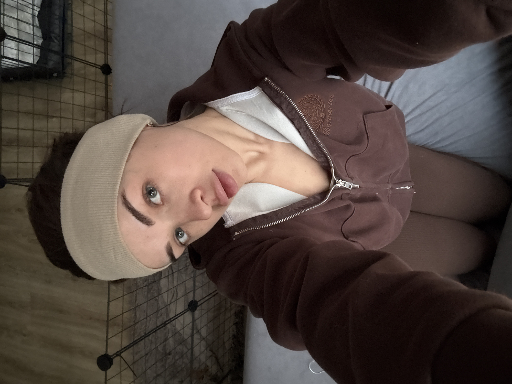 An indoor, overhead selfie features a person with fair skin, light eyes, and dark eyebrows, wearing a light-colored headband that covers their forehead and ears. Their dark hair is pulled back. The person is looking slightly upwards and to the side, with their tongue playfully protruding. They are dressed in a partially unzipped brown zip-up hoodie over a white undershirt. The hoodie has an embroidered logo on the left chest area, featuring a crest design and the text "Bonnerbee" clearly visible beneath it. The zipper is silver-toned.
The background shows a grid-like metal structure on the left side, possibly a pet enclosure or modular shelving, with a light wooden floor visible beneath and through it. A dark object, possibly bedding or a bag, is inside the enclosure. To the right and beneath the person, a light gray fabric surface, likely a bed or sofa, is visible. The overall setting suggests a casual, possibly home, environment. The image is rotated 90 degrees clockwise from a typical upright orientation. The general lighting is soft, indicating an indoor, likely daytime, setting.