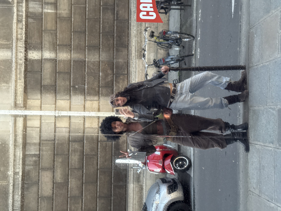 A mirror selfie captures two individuals standing on a tiled sidewalk of a street in Paris, France. Visible water droplets are present on the reflective surface.
The individual on the left has dark skin and an Afro hairstyle, wearing a cropped brown jacket over a dark crop top, baggy brown pants, and black boots. Their right hand is raised, displaying a peace sign. A multi-layered belt with pouches is worn at the waist.
The individual on the right has lighter skin, long dark hair covered by a patterned, studded cap, and wears a grey jacket over a light crop top, baggy light-wash jeans, and dark brown boots. This individual holds a silver smartphone in their right hand, aimed to capture the image. Their left hand rests on a dark, studded pole.
In the reflected background, an asphalt street is visible. Parked vehicles include a partial silver car on the far left, a bright red Vespa-style scooter behind the left individual, and several bicycles further right. The background building facade is constructed of large, light-colored stone blocks, with a vertical white pipe running down its center. A red and white sign displaying the word "CAFÉ" is partially visible on the far right of the reflection.