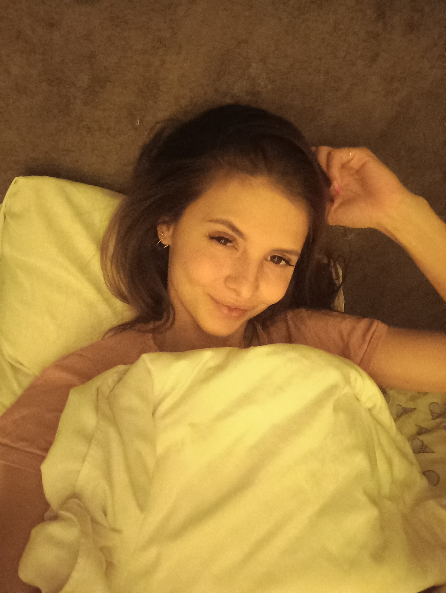A single female subject is depicted lying in bed within an indoor setting, likely a bedroom, in the city of Unknown, Unknown. The subject, appearing to be in her 20s or early 30s, possesses long dark brown hair, fair skin, and is wearing a light pink short-sleeved t-shirt. Her head rests on a light green/yellowish pillow. Her right arm is bent with the hand positioned near her head, lightly touching her hair; a thin, light-colored ring is visible on the ring finger of her right hand, and her fingernails exhibit pink polish. She is covered from the chest down by a light yellow/cream-colored duvet or blanket. Beneath the duvet on the right, a light green/yellowish sheet with faint, light-colored patterns is visible. The background above her head and to her right consists of a dark brown, textured surface, possibly a headboard or wall. The subject maintains direct eye contact with the viewer, exhibiting a subtle, closed-mouth smile. Her left ear displays two earrings: a small stud and a small hoop. The scene is illuminated by warm, yellowish light, suggesting an ambient or artificial light source.