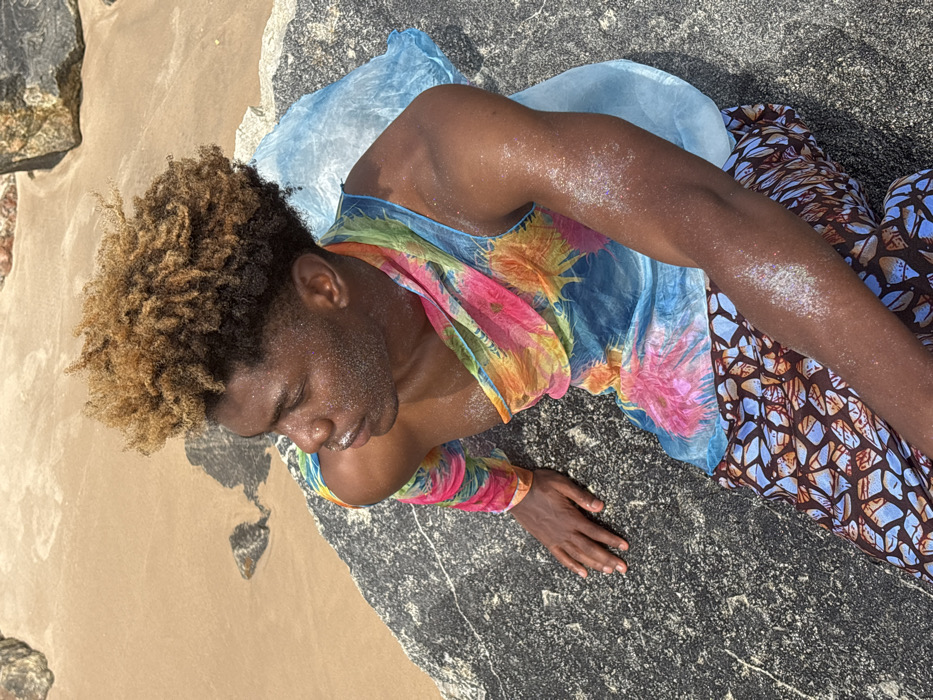 A male individual with dark skin and short, bleached blonde-brown curly hair is positioned on a large, dark, textured rock at a beach in Alaguntan, Nigeria. He is seated, looking towards the left side of the frame with his head slightly tilted. His face and exposed arms are speckled with reflective glitter or shimmering particles. He wears a sheer, multi-colored garment featuring a tie-dye or abstract pattern in shades of blue, pink, orange, and yellow, with one long sleeve covering his left arm and the fabric draped across his torso. Below, he wears lower attire with a brown and blue geometric pattern. A light blue, translucent fabric or object is visible behind and to his right, resting on the rock. The immediate background consists of light brown sand with additional dark rocks, indicating a coastal environment under bright sunlight.