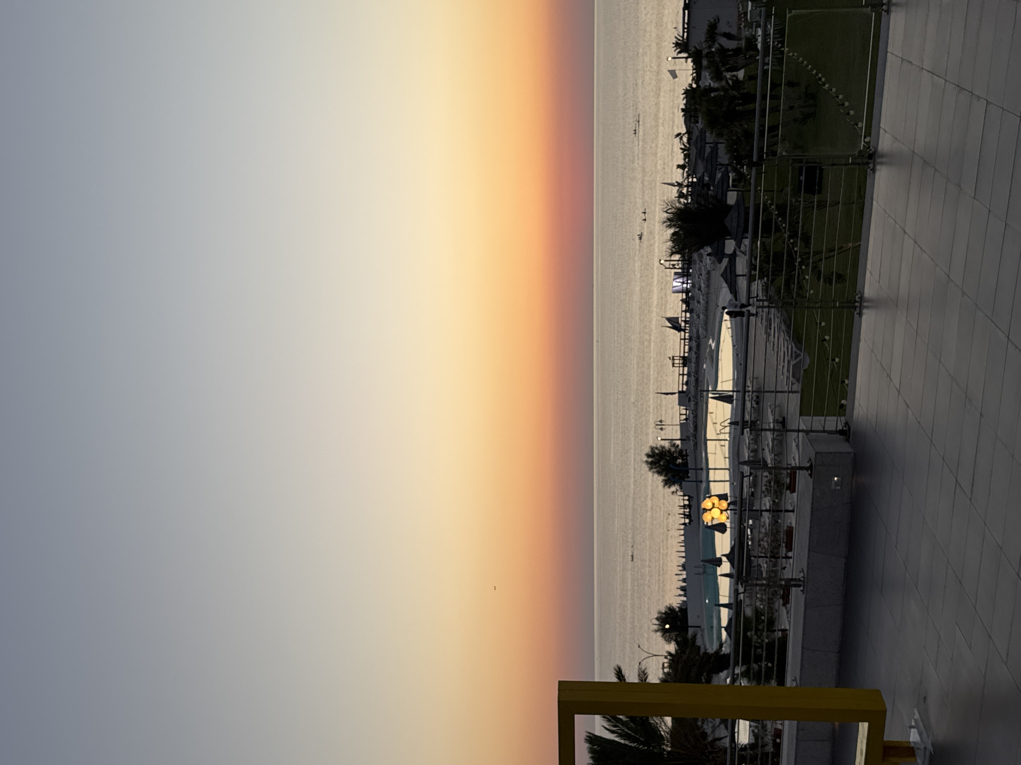 The image captures a sunset over the sea in Isola delle Femmine, Italy. A vast expanse of ocean meets a gradient sky of pale blue fading into warm orange and pink hues. In the foreground, a tiled patio area overlooks a resort-like setting. Beyond the patio, a swimming pool is visible, surrounded by lounge chairs and umbrellas. Palm trees and other vegetation dot the landscape. The water appears calm, with a few small figures in boats visible in the distance. A yellow structure is partially visible on the left edge of the frame. The overall scene depicts a tranquil evening at a seaside resort.