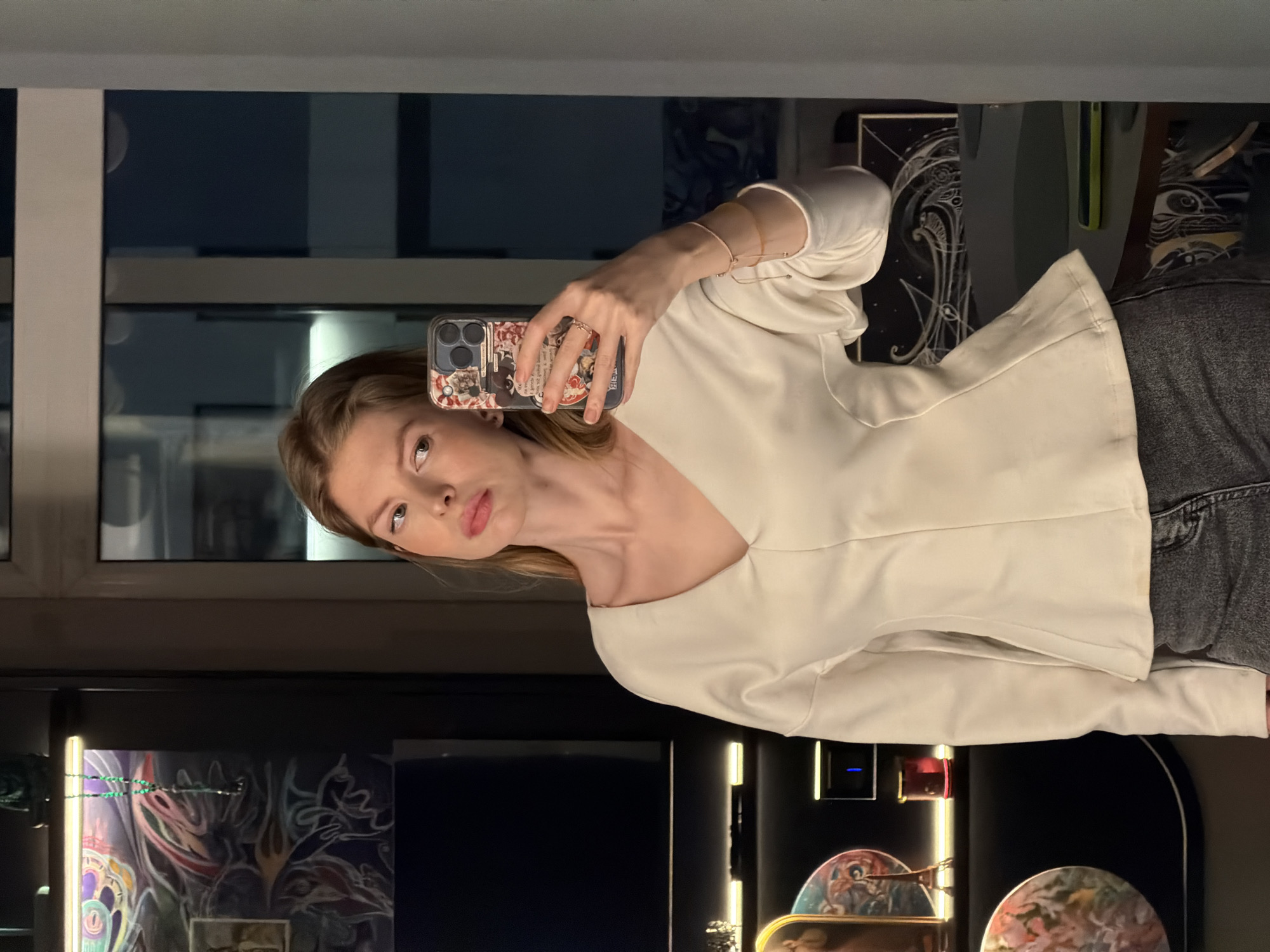A fair-skinned female is taking a selfie in what appears to be an upscale interior space. She is holding a smartphone with a decorative case, positioned in front of her face. She is wearing a white v-neck blouse and what seems to be a dark denim garment. The setting features a large window revealing an urban cityscape, likely Moscow, Russia, judging by architectural styles and typical light conditions. Below the window, there's a reflective surface displaying various objects, including decorative items with painted figures. Artificial lighting enhances the display. The focus is sharply on the woman and her immediate surroundings.