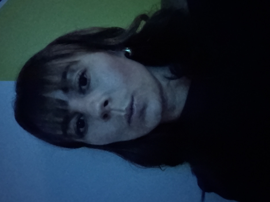 A woman with shoulder-length dark hair and bangs looks at the camera. She is fair-skinned and wearing a dark shirt. There appears to be a dark earring. The background is divided into three distinct sections: a blue area at the bottom left, a green area at the top left, and a dark area at the right. The lighting is dim, with a bluish tint overall.