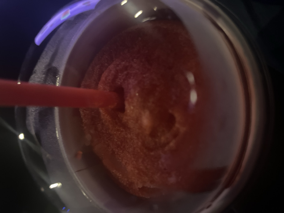 A close-up image depicts a red, frozen, slushy-textured beverage contained within a clear plastic cup with a domed lid. A red plastic straw is centrally inserted through the lid, extending into the drink. The beverage exhibits a granular, icy consistency and a uniform red coloration. The background is dark and blurred, featuring indistinct light reflections, some with a purple hue, suggesting an indoor, low-light environment. The scene is located in Mexico City, Mexico. No individuals or specific actions beyond the presence of the beverage and straw are discernible.