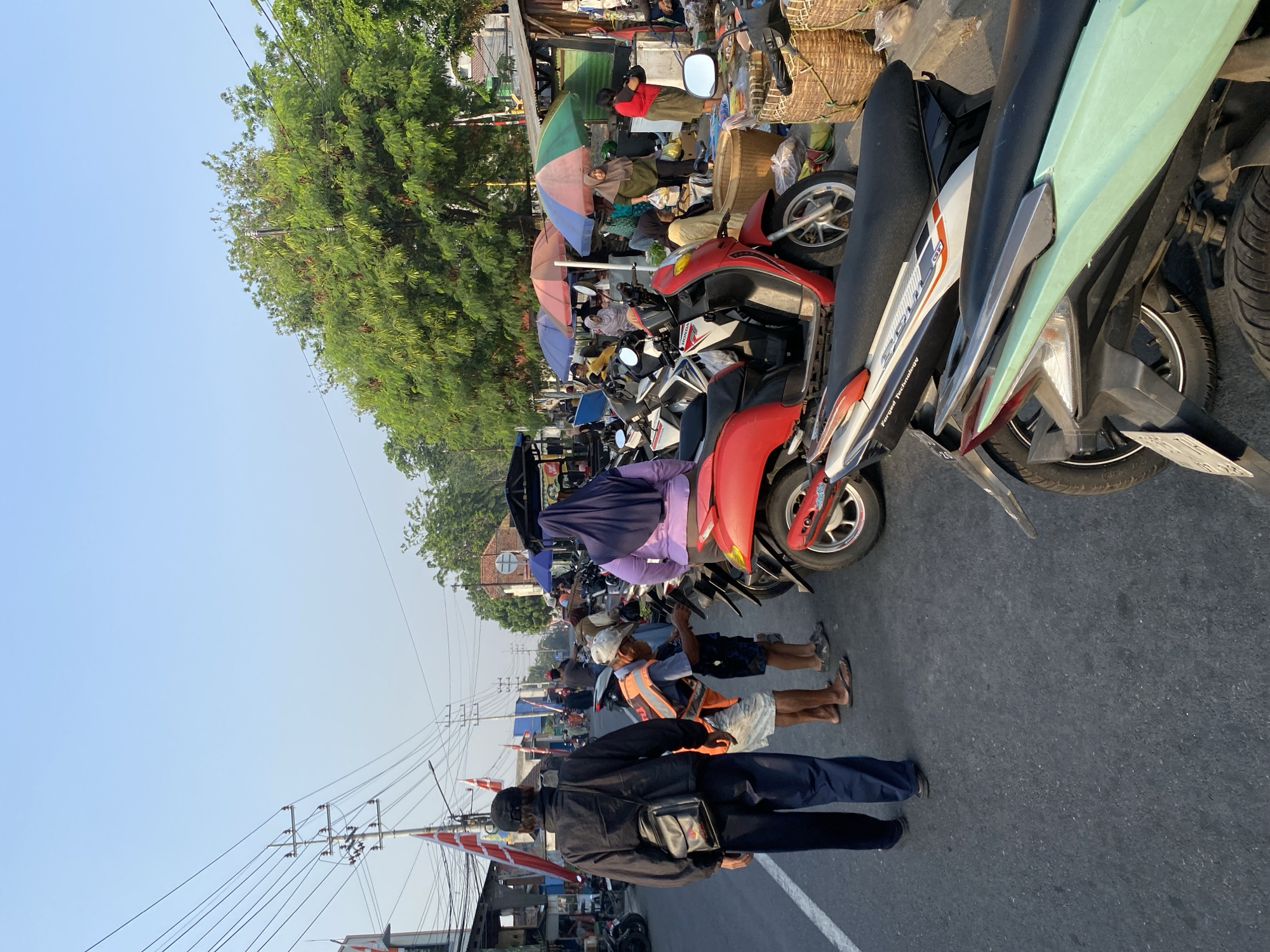 The image depicts a busy street scene in Tebel, Indonesia, likely a market area, bathed in the soft light of early morning or late afternoon. The street is lined with parked motorcycles and various stalls, suggesting a marketplace or a commercial district. Power lines crisscross overhead, typical of urban environments in Indonesia.
In the foreground, the back of a person wearing a black jacket and dark pants, with a black shoulder bag, is visible, facing away from the camera. To their right, two individuals are interacting with a parked motorcycle. One, wearing an orange safety vest and a cap, leans over the motorcycle, possibly attending to it or speaking with someone. The other person, a young boy in shorts, stands nearby.
Further into the scene, a woman in a purple hijab stands near a red motorcycle. A colorful array of market stalls with umbrellas provide shade to vendors and shoppers. People are seen browsing or engaged in transactions. Baskets filled with goods are stacked near some of the stalls, hinting at the types of products being sold, which could range from food to household items. The presence of multiple motorcycles indicates common modes of transportation and the general atmosphere of a vibrant community gathering or marketplace. The overall environment is an active, outdoor commercial space characterized by a mix of vehicles, people, and market infrastructure under a clear sky.