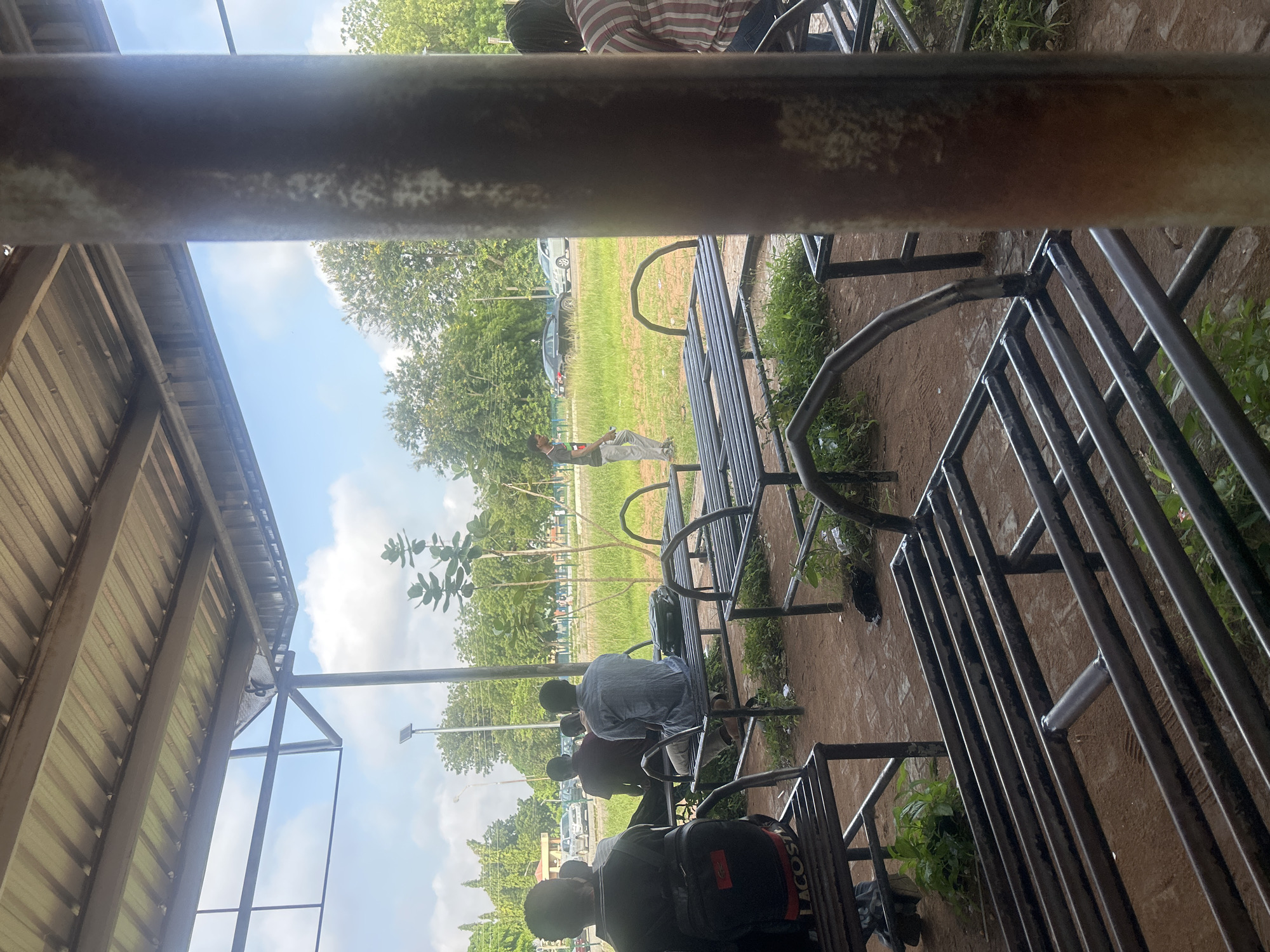 An outdoor seating area in Ogbomosho, Nigeria, features several metal benches and tables under a corrugated metal roof supported by pillars and metal beams. The ground is a reddish-brown dirt, with some grass and weeds growing around the seating. In the background, trees and greenery are visible, along with a road where cars are parked and driving. Several people are seated on the benches, facing away from the camera, and one person is standing and walking. The sky is partly cloudy.