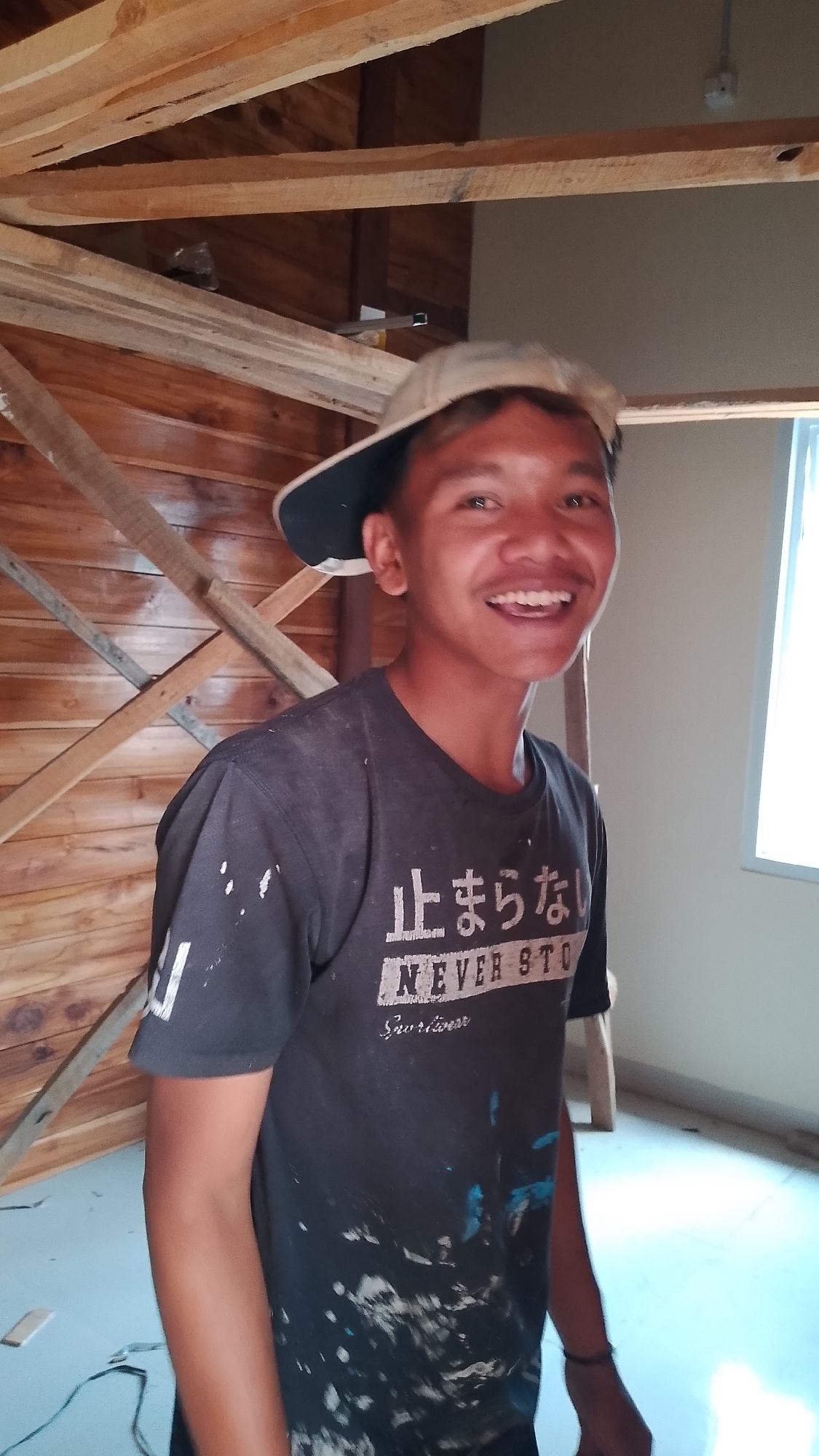In Karanglangu, Indonesia, a young male, appearing to be in his late teens or early twenties, stands within a room under construction. He wears a dark t-shirt splattered with white and blue paint, a beige baseball cap worn backward, and a black bracelet on his left wrist. The t-shirt features Japanese characters and the text "NEVER STO" above "Survivor". He is smiling broadly, with his mouth open, exposing his teeth. The background reveals unfinished wooden walls and ceiling beams, characteristic of a construction site. To the right, a partially visible window allows natural light into the space. The floor appears to be a light-colored material, possibly concrete or a similar finishing. The overall impression is of a worker taking a brief moment for a photograph amidst ongoing construction.