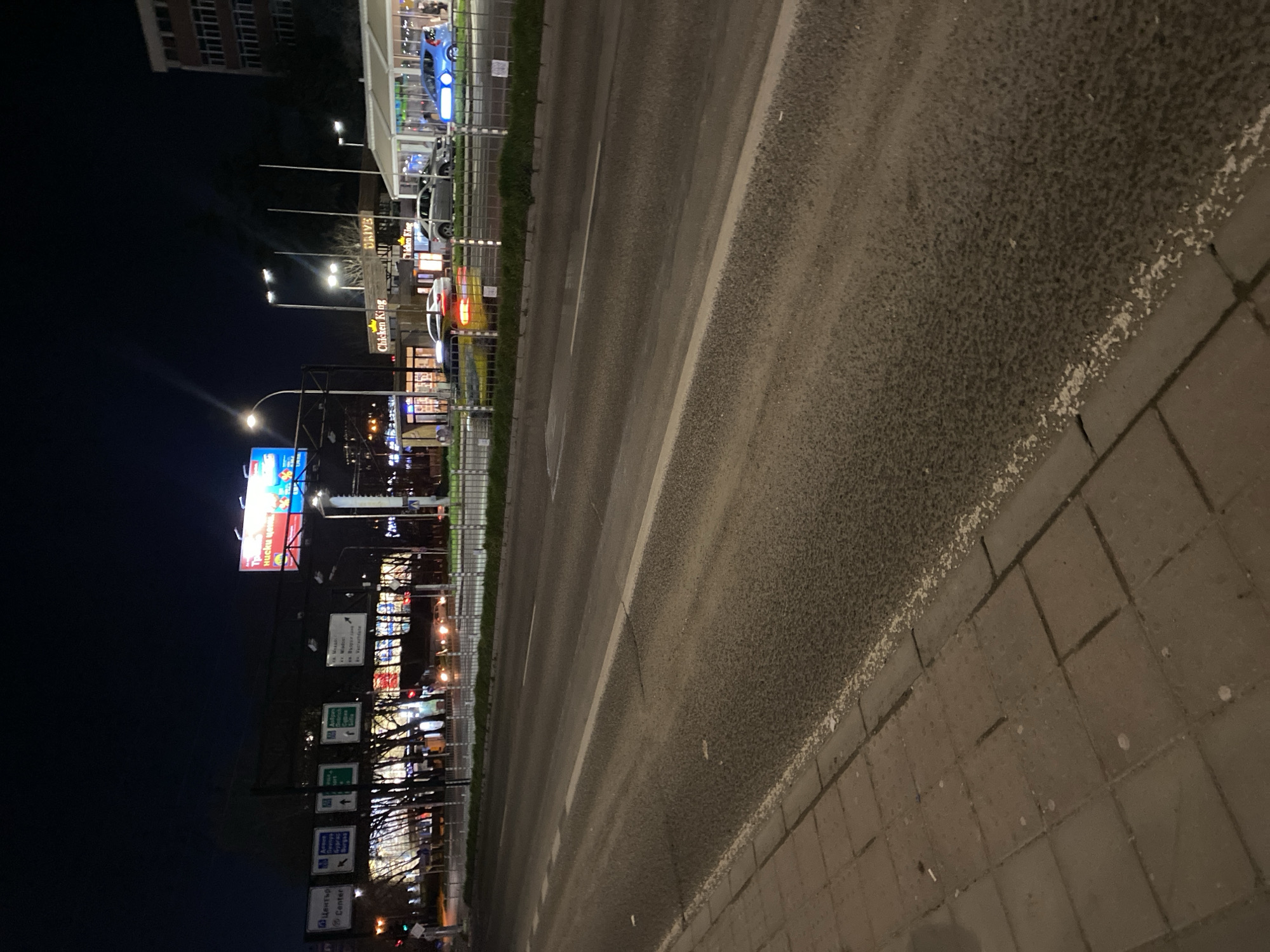 The image depicts a vibrant urban street scene at night in Varna, Bulgaria. The central feature is a multi-lane asphalt road, illuminated by streetlights, with clear white dashed and solid lane markings. To the right, a sidewalk made of reddish-brown square pavers runs alongside the road. The sky is dark, indicating late evening or night.
On the left side of the road, a commercial strip is brightly lit with numerous signs and building lights. A prominent sign for "Chicken King" is visible, along with a "DRIVE" sign, suggesting a fast-food drive-thru. Several cars, including a blue vehicle and others with illuminated red and yellow taillights, are visible near these establishments. Further back on the left, a tall building with many windows adds to the urban backdrop.
A series of road signs are placed along the left side of the street. One sign clearly indicates "Център" (Center) in Cyrillic and "Center" in English, accompanied by a parking symbol. Other signs, predominantly in Cyrillic, appear to direct traffic towards destinations such as "Варна" (Varna), "Бургас" (Burgas), and potentially "Варненски Университет" (Varna University) and "ОБЛАСТНА УПРАВА" (Regional Administration). A large illuminated billboard displays Cyrillic text, possibly "НИСКИ ЦЕНИ" (Low Prices). The area is separated from the road by a metal fence. The scene captures the active atmosphere of a commercial district after dark.