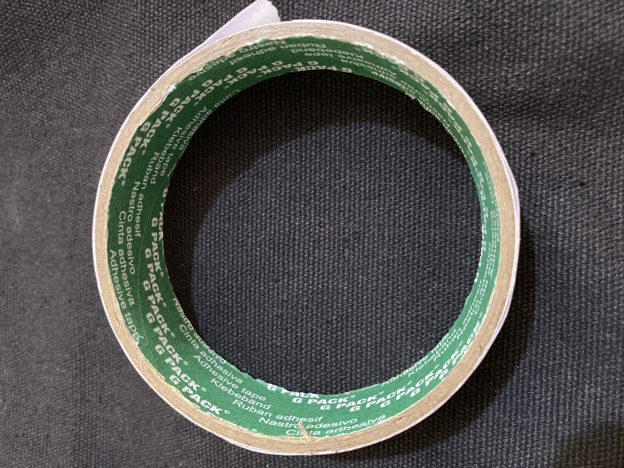 A roll of green and white adhesive tape, branded with "G PACK" and various translations of "adhesive tape" like "Cinta Adhesiva," "Nastro Adesivo," "Ruban Adhesif," and "Klebeband," is shown from a top-down perspective. The tape is laid flat on a dark, textured fabric surface, possibly a dark gray or black cloth. The roll is circular, with the inner core slightly visible. The lighting suggests an indoor environment with a light source positioned above and to the right of the roll, casting subtle shadows. There are no people or animals present in the image, and the context does not provide any information about the city of Lhokseumawe, Indonesia. The scene is static and focuses solely on the object.