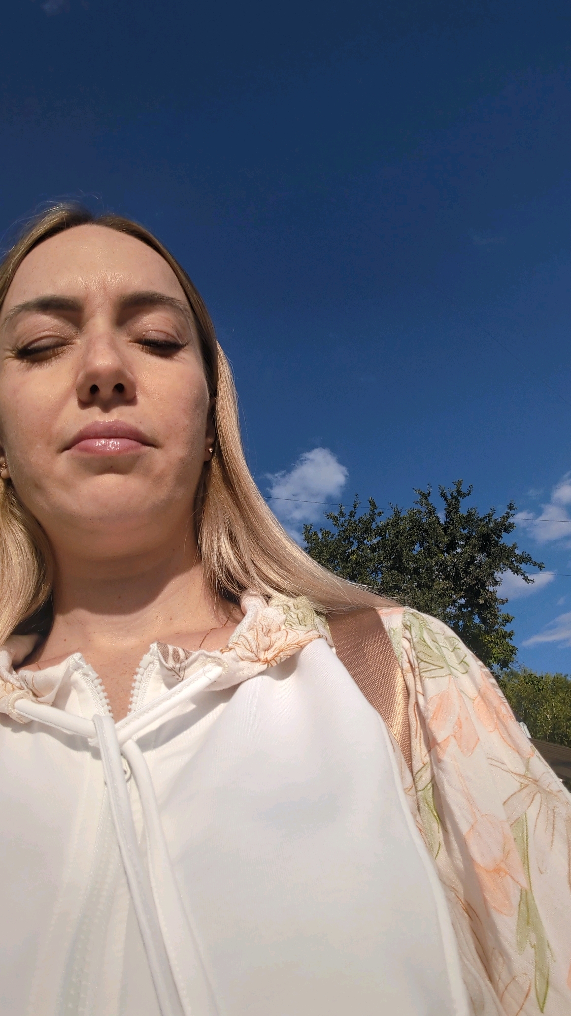 A young woman with blonde hair and closed eyes is photographed outdoors, looking upwards against a bright blue sky with scattered clouds. She is wearing a white zip-up jacket over a floral-patterned garment. A tree with dark green foliage is visible in the background, to the right of the woman. The image does not provide sufficient information to determine the location within Chișinău, Moldova.