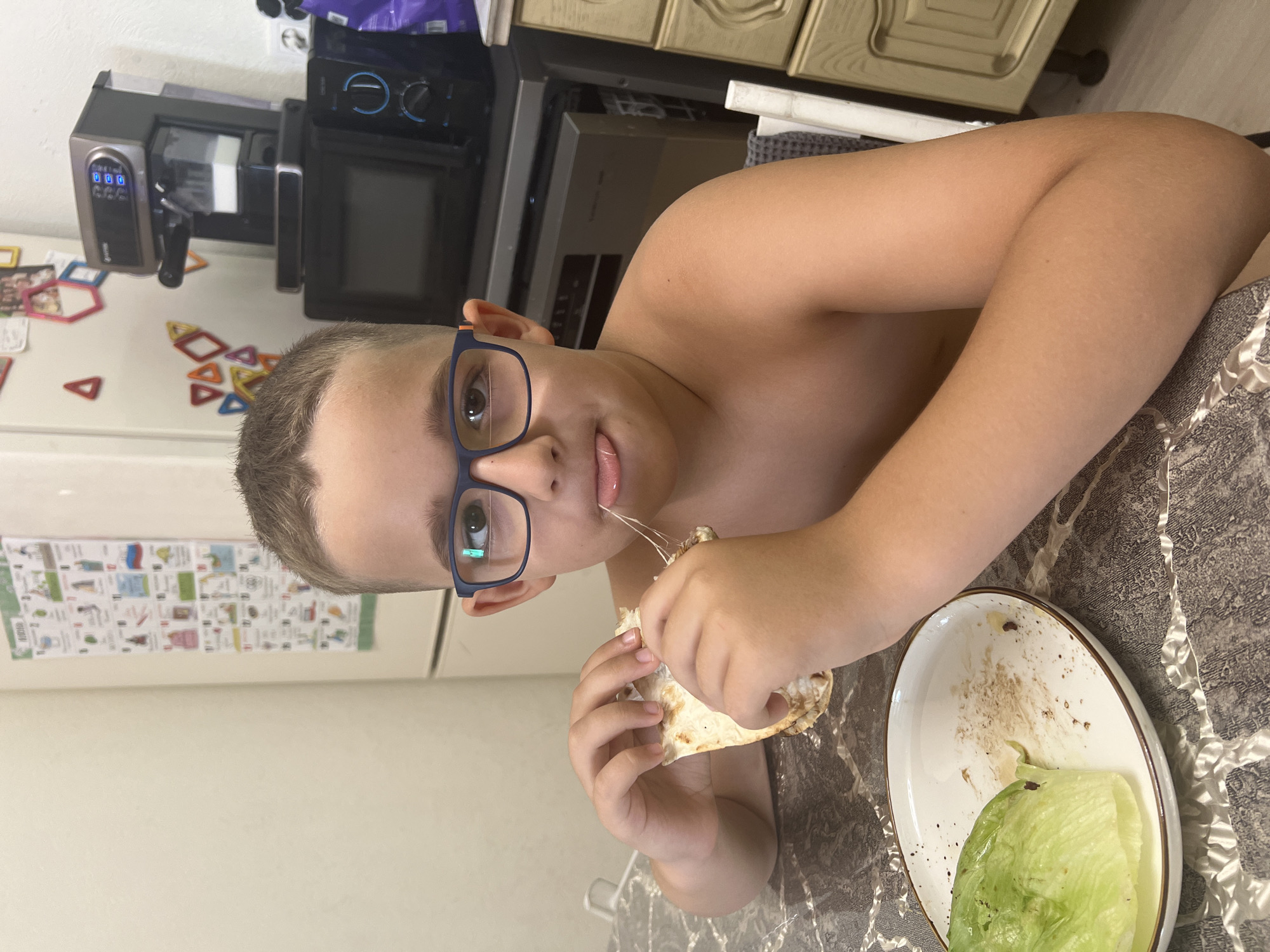 A young boy with short, light brown hair and blue-framed glasses is shown in a close-up, horizontal shot, looking directly at the camera with a faint smile. He is shirtless and appears to be seated at a table covered with a patterned, possibly textured, tablecloth. He holds a piece of flatbread, likely a wrap or quesadilla, with both hands, actively taking a bite from which long, melted cheese strings stretch from his mouth to the food.
To his right, a white plate with a brown rim sits on the table, containing a large piece of green lettuce and some food remnants.
The background reveals a kitchen setting. A black microwave oven is visible behind the boy's head. Next to it, a silver and black appliance, possibly a coffee maker or a water dispenser, displays "000" on a digital screen and features a logo that appears to be "GETTO". Above these appliances are light-colored wooden cabinets with decorative carvings. On a white refrigerator door to the left, numerous colorful magnetic shapes, including squares, triangles, and hexagons, are arranged. A white chart or poster is also affixed to the refrigerator, displaying text that includes the Cyrillic word "ГОЛОНН" on its left side.
The image captures a casual, candid moment of the boy eating, likely at home in Sochi, Russia.